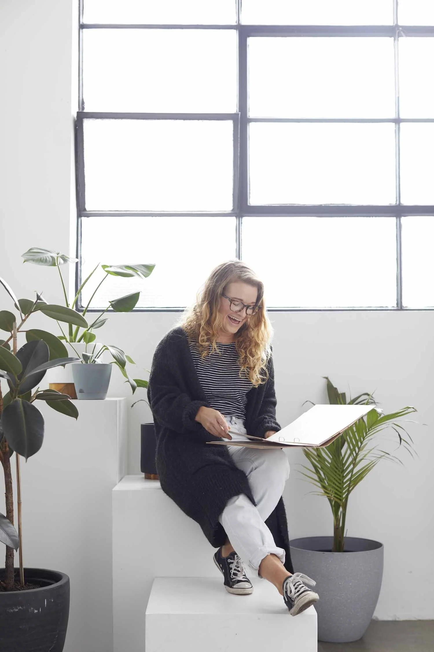 A woman with curly blonde hair and glasses, smiling and looking at a binder, seated on white steps in a bright room with large window and potted plants.