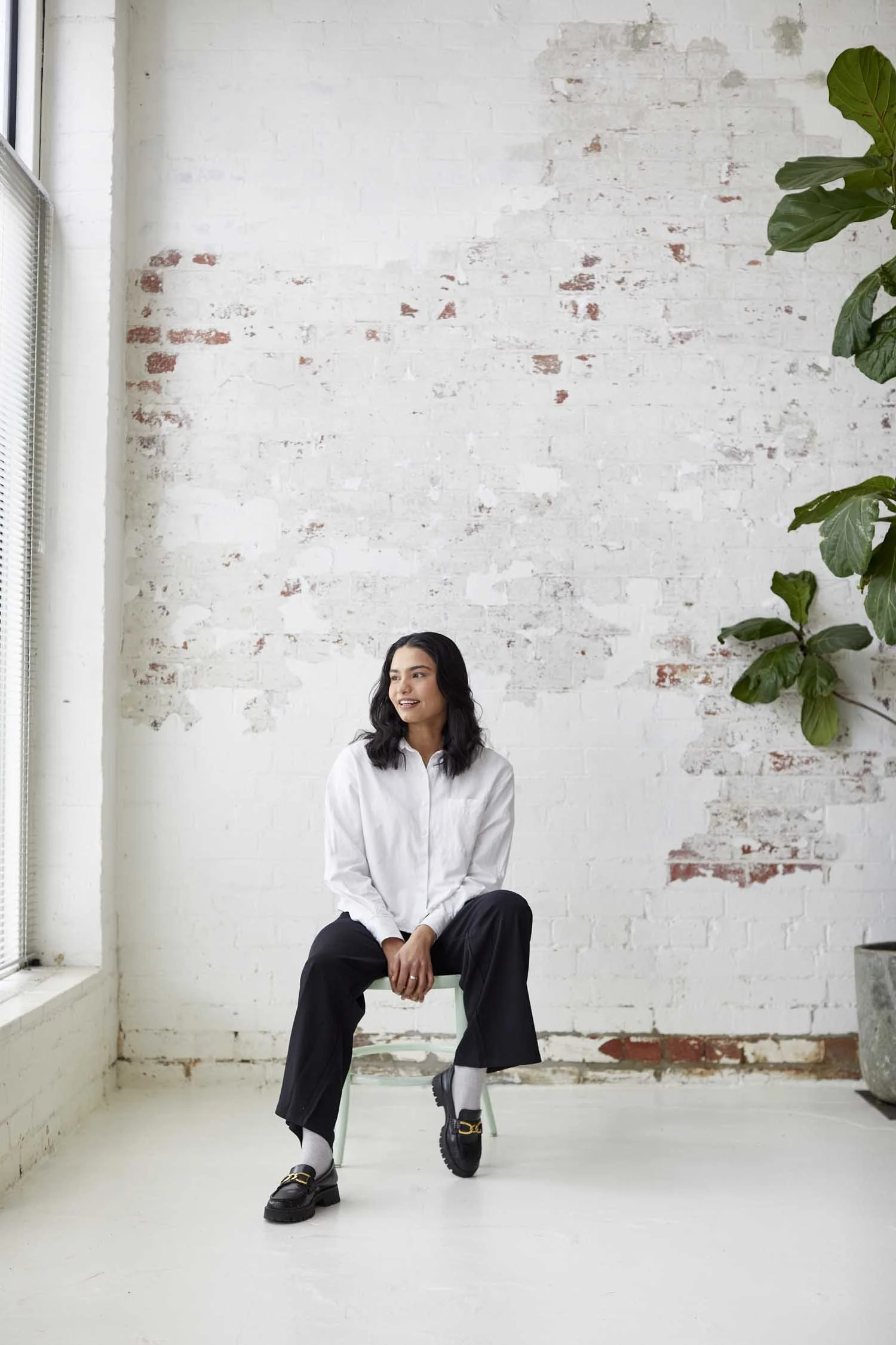 A woman with long black hair sitting on a small chair in front of a white brick wall, wearing a white shirt, black wide-leg pants, chunky black shoes, white socks, and smiling while looking to her right. There is a large green plant on the right side
