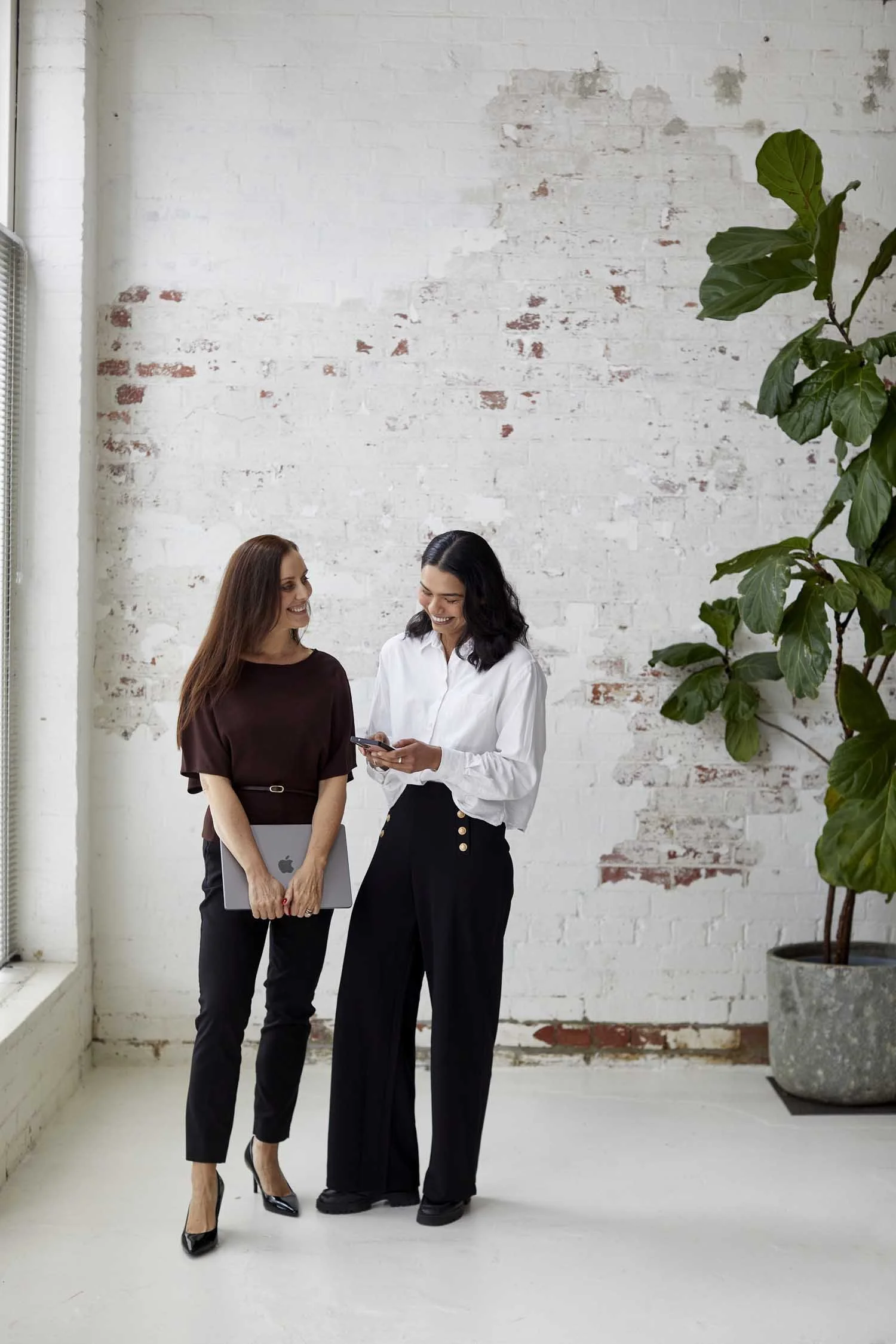 Two women smiling and talking in a bright room with a white brick wall, one holding a laptop and the other holding a phone, with a large green plant on the side.