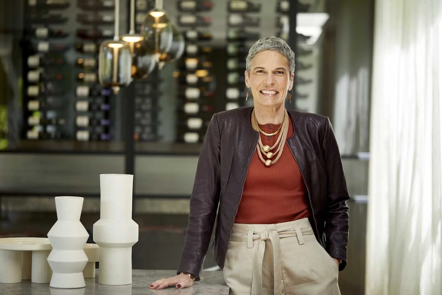A woman with short gray hair smiling in a wine shop or tasting room, standing with her hand resting on a table with decorative white vases, with wine bottles displayed on wall-mounted racks in the background, and sunlight coming in through curtains.