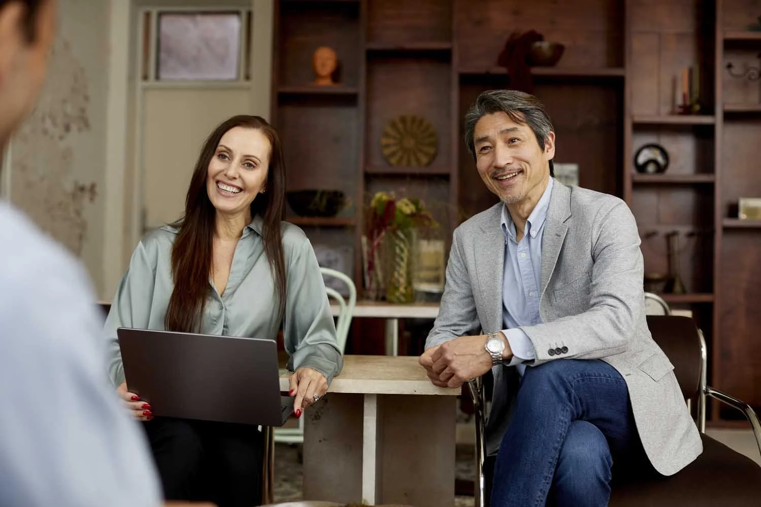 Two adults, a woman with long brown hair and a man with short dark hair, sitting at a table, smiling and engaging in conversation with a person outside the frame in an indoor setting with shelves and decorations in the background.
