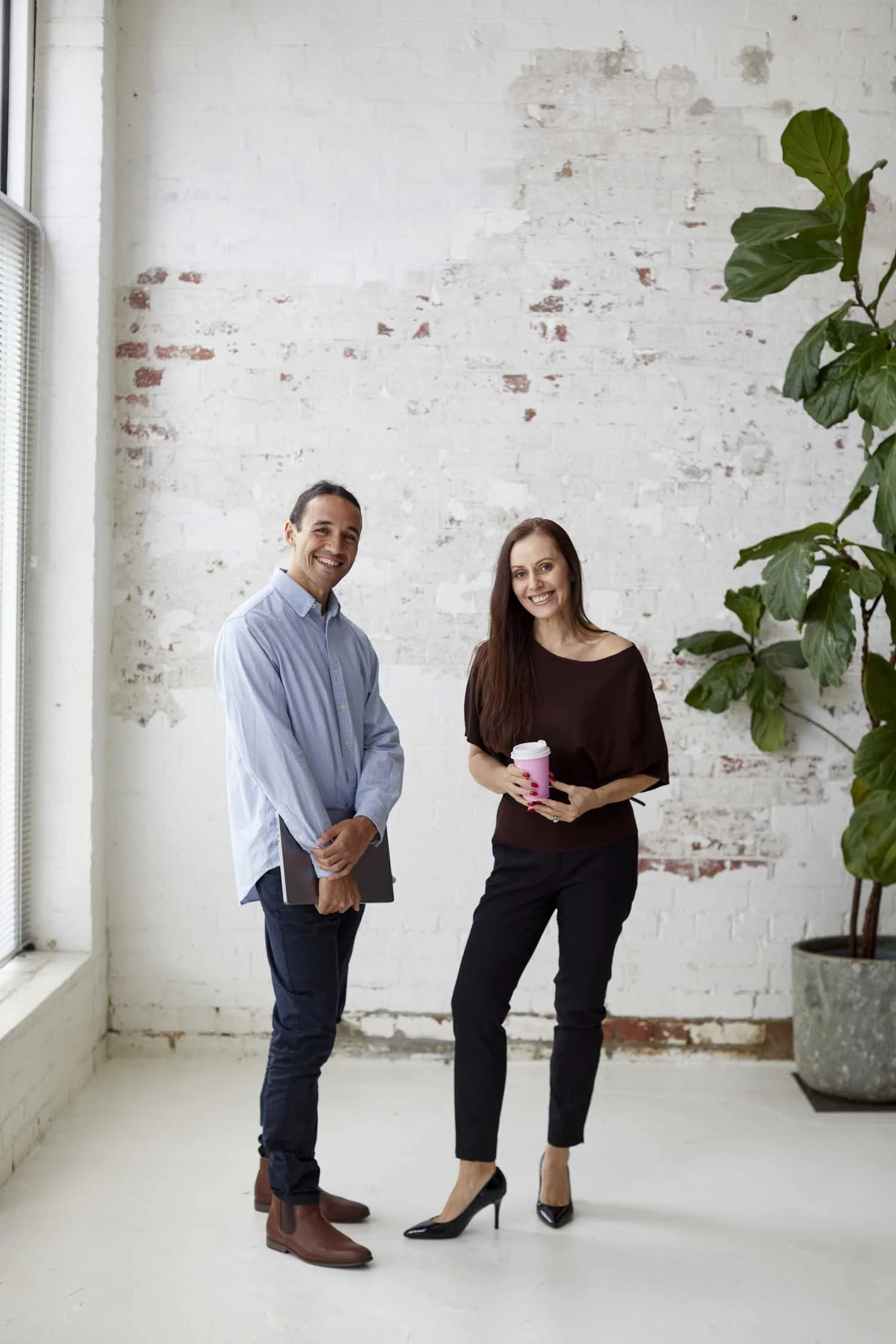 Two professionally dressed people, a man and a woman, standing in front of a white brick wall with a large potted plant nearby. The man is wearing a light blue shirt, dark trousers, and brown shoes, holding a tablet. The woman is wearing a black dres