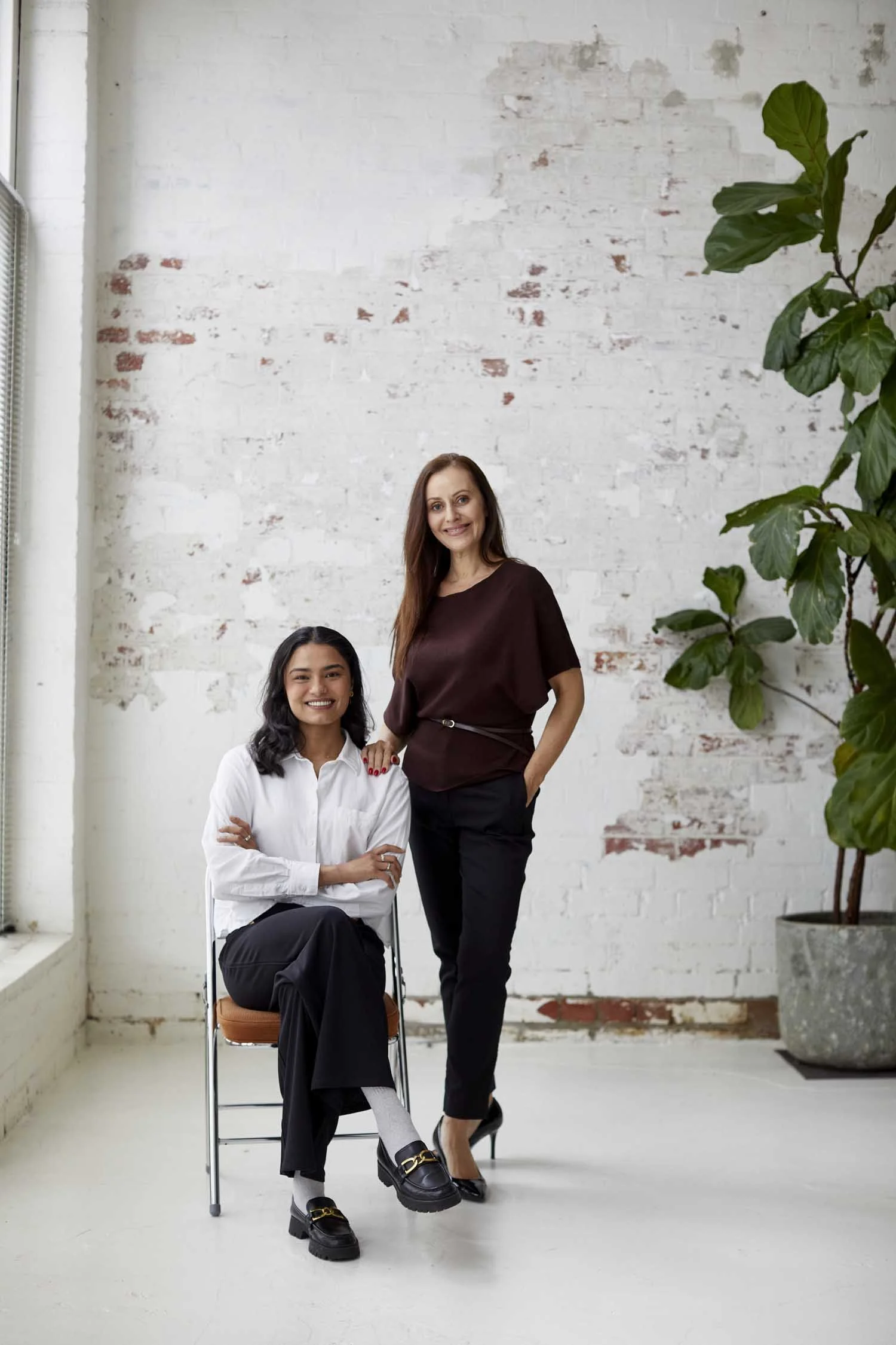 Two women in a bright, modern room with a white, exposed brick wall. One woman is sitting on a chair, smiling with arms crossed, wearing a white shirt and black pants. The other woman is standing beside her, smiling, with one hand on the seated woman