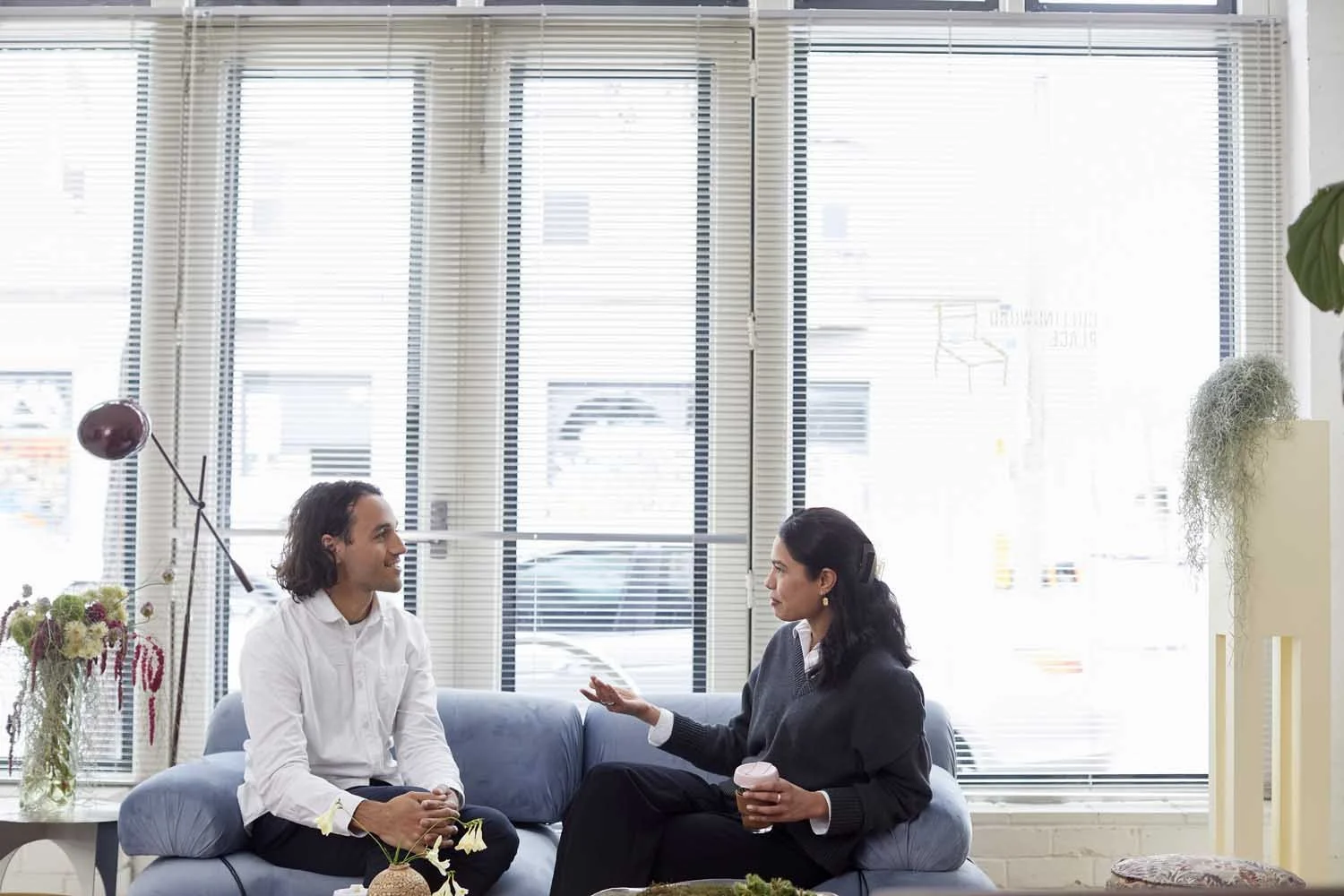 Two people, a man and a woman, sitting on a blue sofa in a bright room having a conversation. The man has long dark hair and is wearing a white shirt, while the woman has dark hair tied back and is wearing a dark sweater. The woman is holding a cup o
