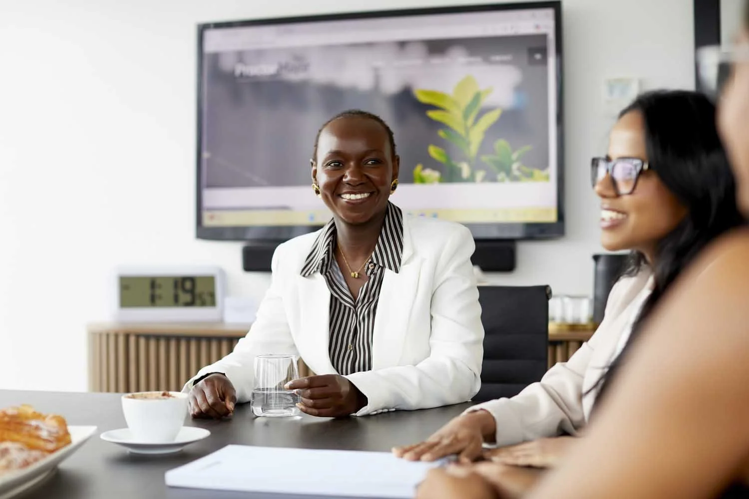 Three women sitting at a conference table, smiling, with a large monitor behind them, in a modern office setting.
