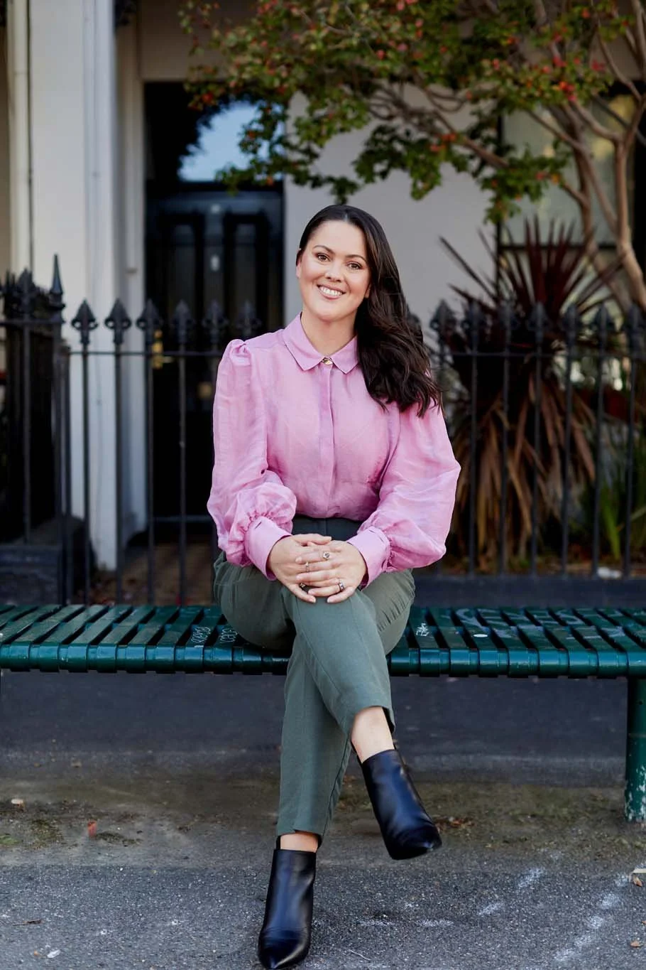 A woman with dark hair, wearing a pink blouse and olive green pants, sitting on a green metal park bench in front of a black fence and house, with trees and plants in the background.