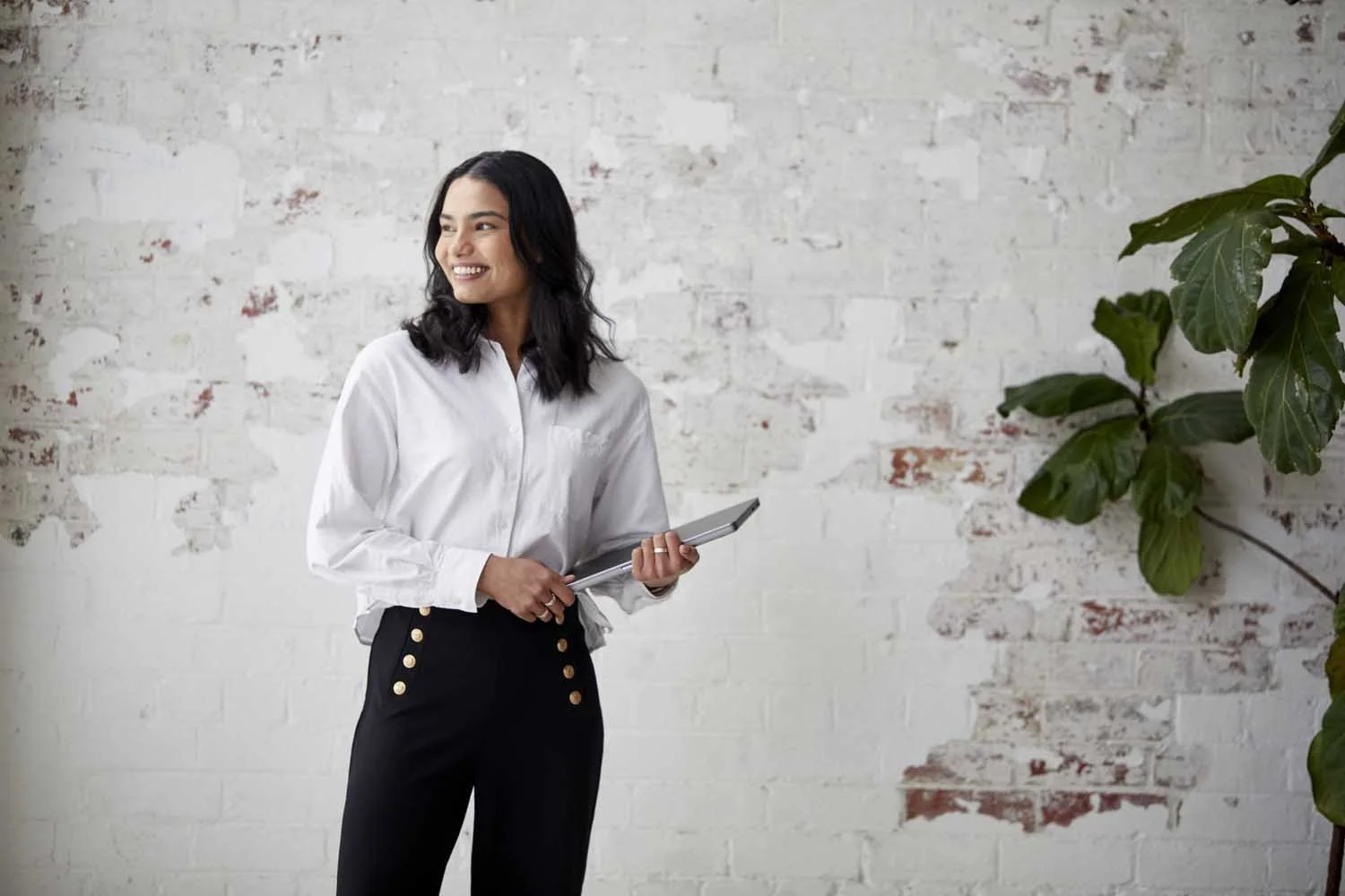 A woman with dark hair in a white shirt and black pants holding a tablet, standing in front of a white brick wall with a large green plant on the right.