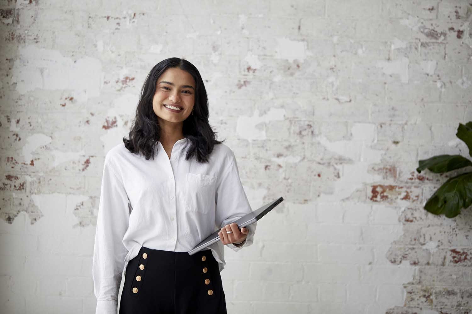A smiling woman with black hair, wearing a white button-up shirt and black pants with gold buttons, standing in front of a white brick wall holding a tablet.