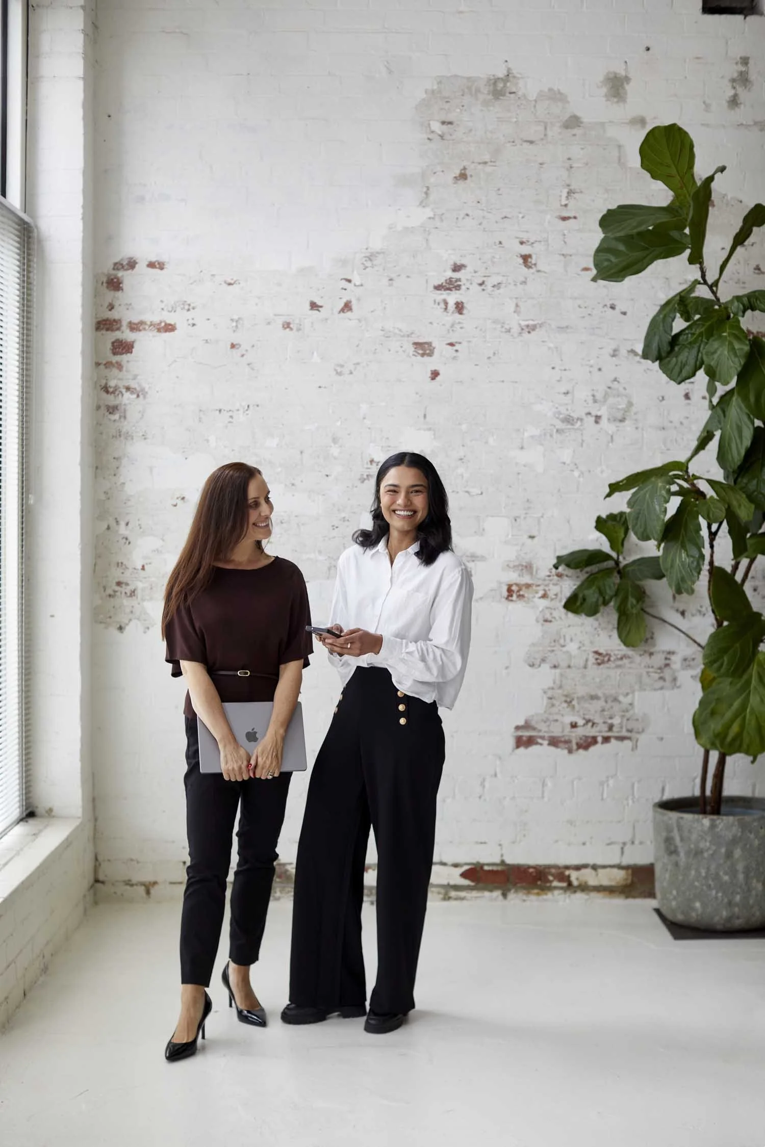 Two women standing in a minimalist industrial-style room with a white brick wall, one holding a laptop and the other holding a smartphone, both smiling and engaged in conversation, with a large leafy indoor plant in a concrete pot to the right.