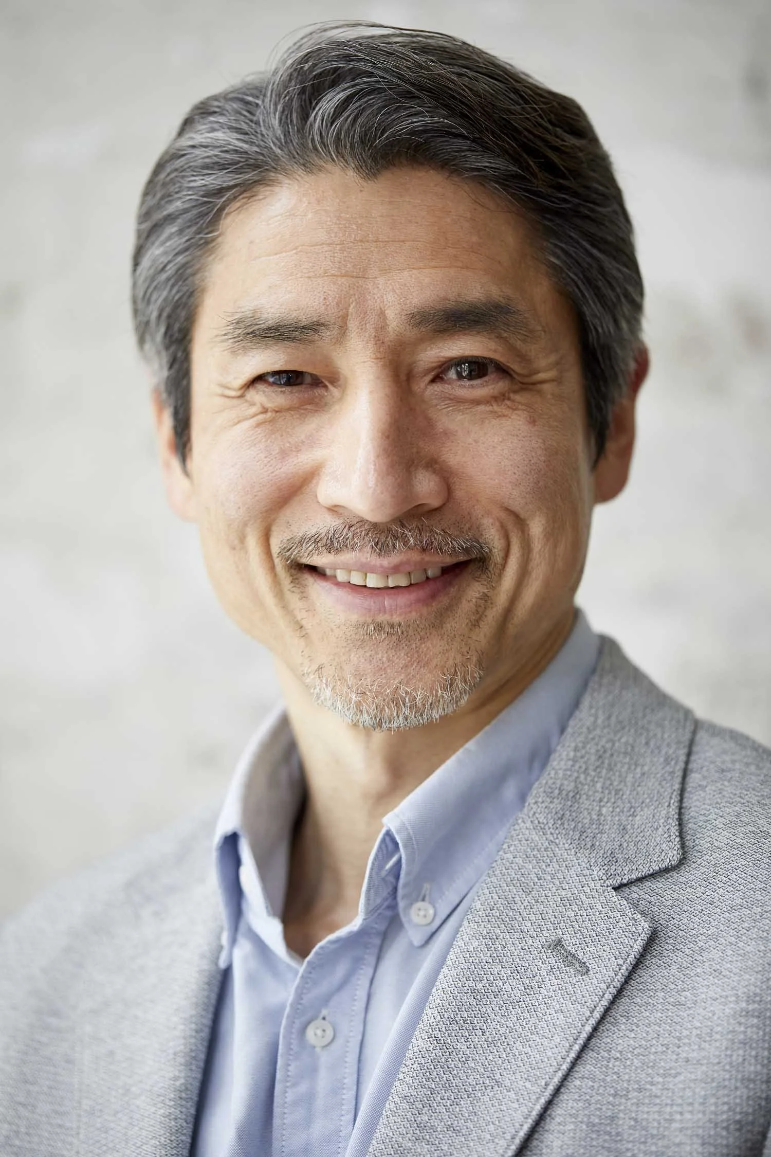 Close-up of a smiling middle-aged Asian man with dark hair streaked with gray, wearing a light gray blazer over a light blue collared shirt, standing against a neutral background.