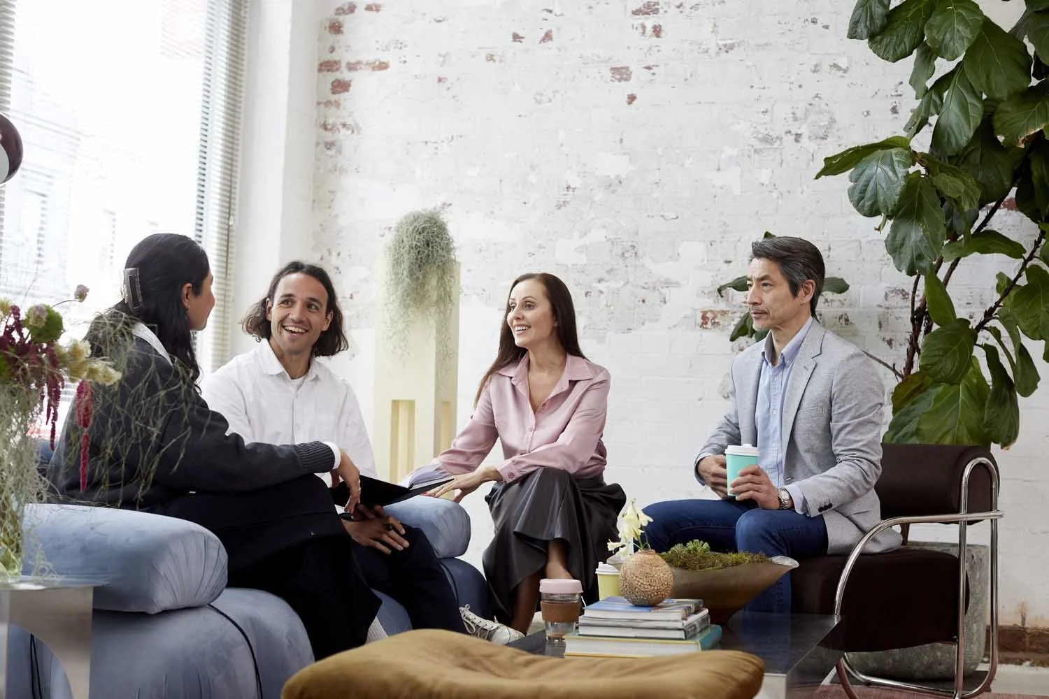 Four people sitting on couches and chairs in a brightly lit modern room, engaged in conversation, with plants and books decorating the space.