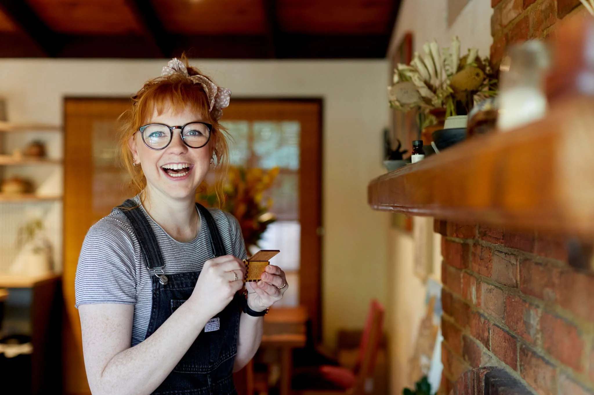 A joyful woman with red hair, glasses, and freckles wearing a striped shirt and black overalls, holding a small wooden object in a cozy home interior with a brick wall and shelves.