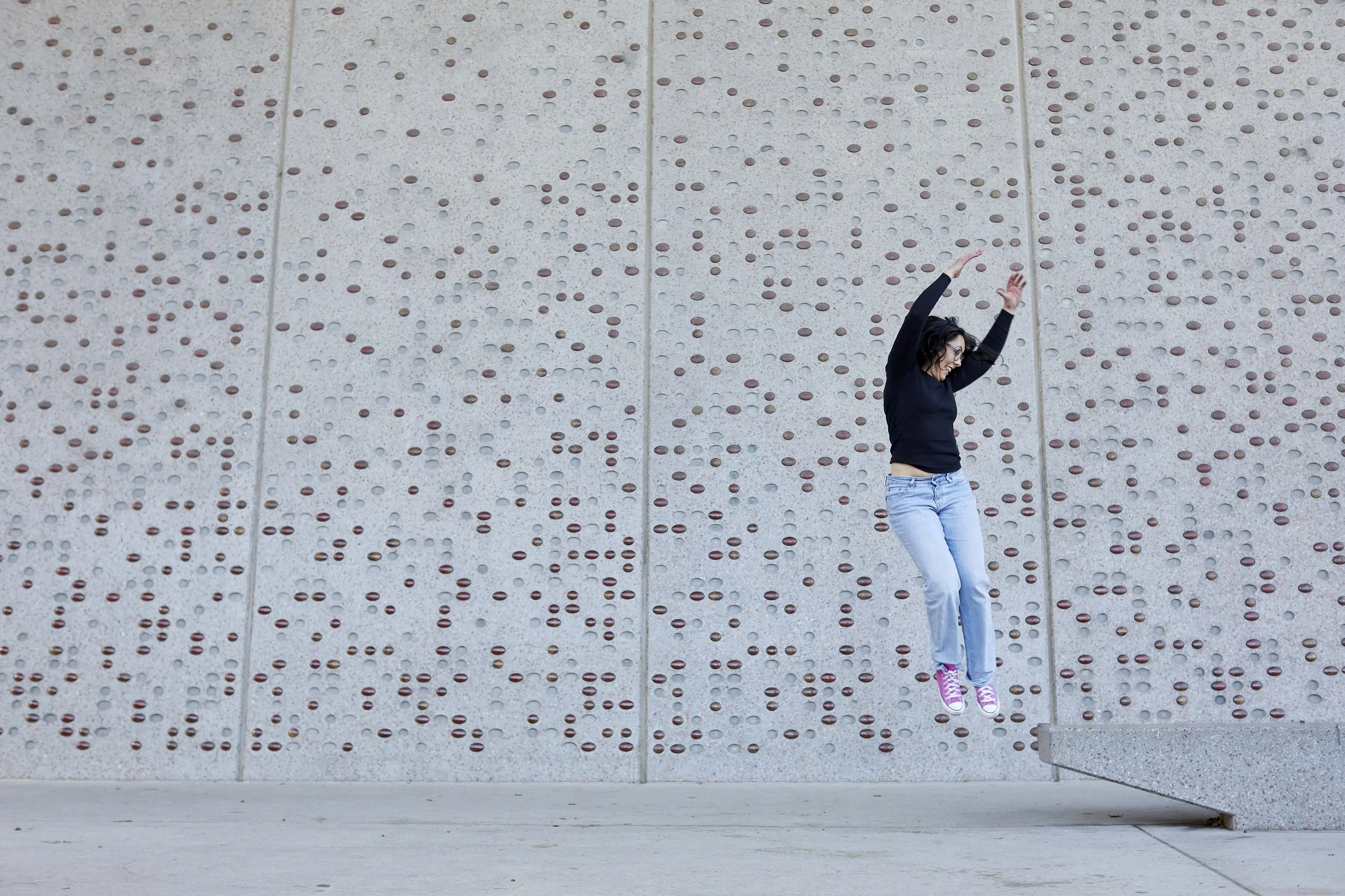 A woman in black shirt and blue jeans jumping with arms raised in front of a concrete wall with circular patterns, wearing pink and white sneakers.