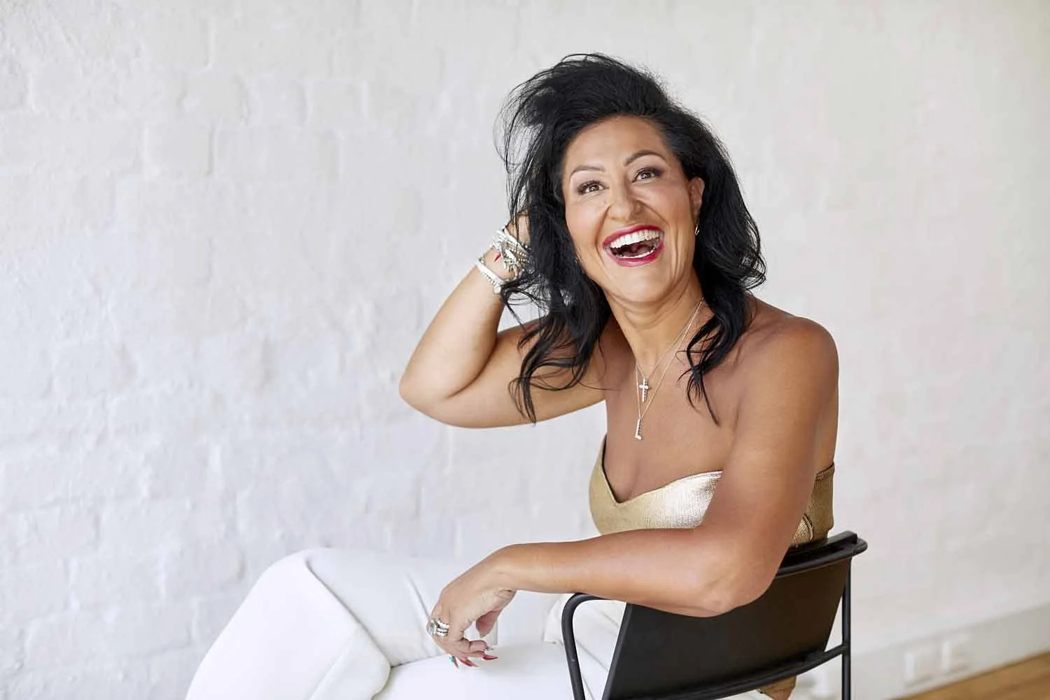 A woman with black wavy hair, wearing jewelry and a strapless top, is seated on a black chair, smiling broadly, with her right hand touching her hair, against a white brick wall background.