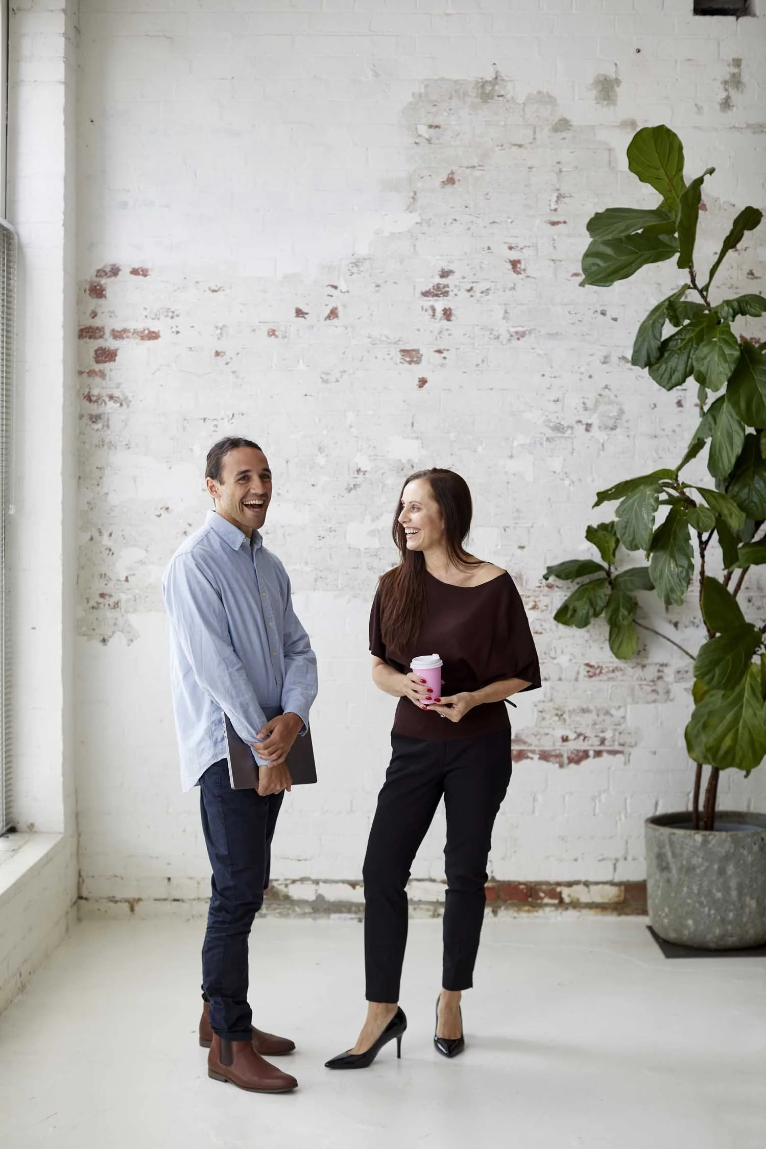 Two professionally dressed people, a man and a woman, are standing and smiling together in an industrial-style room with a white brick wall. The man holds a black folder and wears brown boots, while the woman holds a pink coffee cup and wears black h