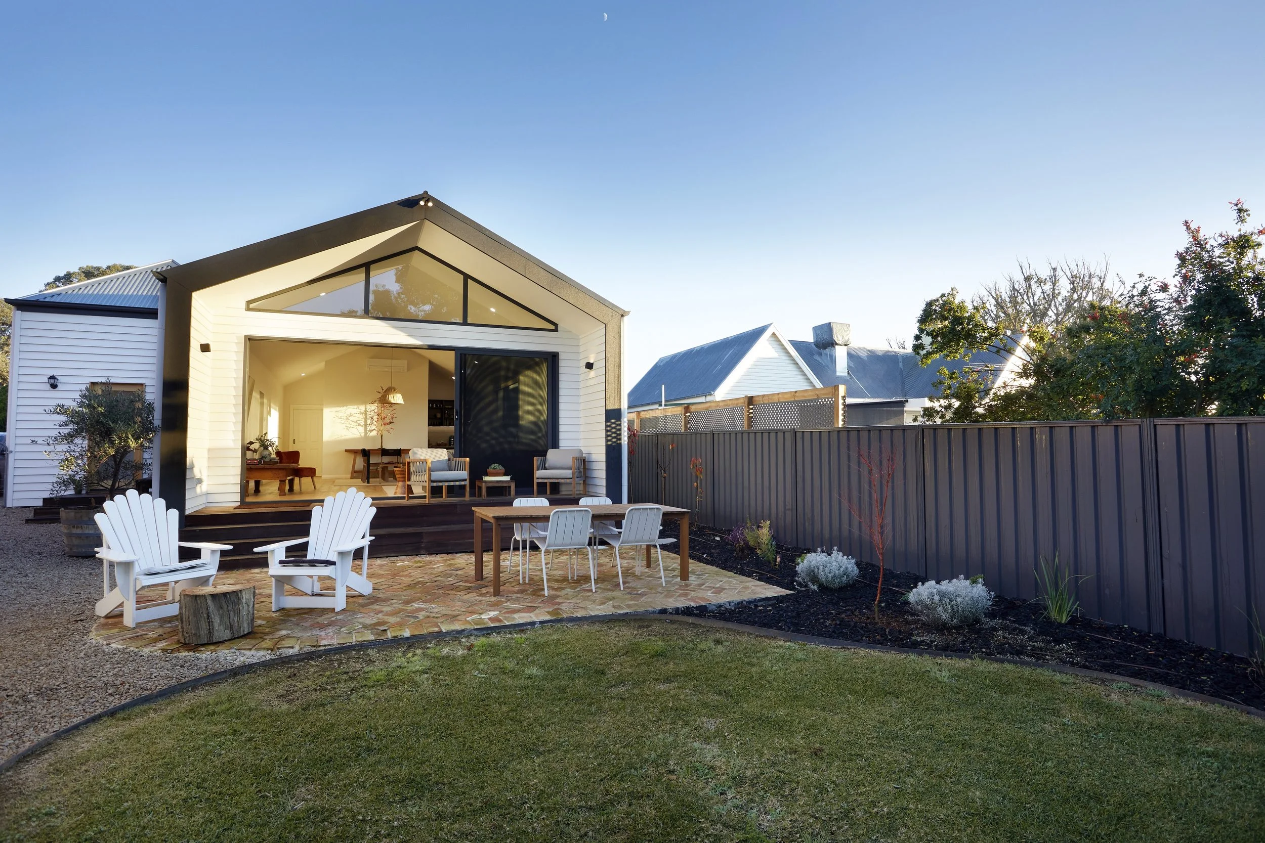 Modern house with large glass doors open to backyard patio, outdoor seating area with white Adirondack chairs, brick paving, garden with plants, and a fenced yard under a clear blue sky with a crescent moon.