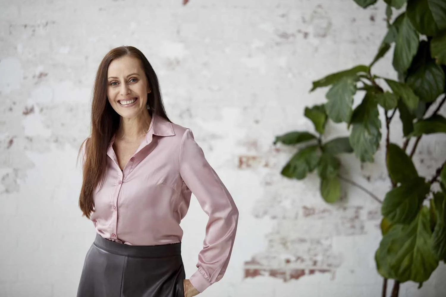 A woman with long brown hair, wearing a pink satin blouse and a dark gray skirt, standing in front of a white brick wall and green plant, smiling at the camera.