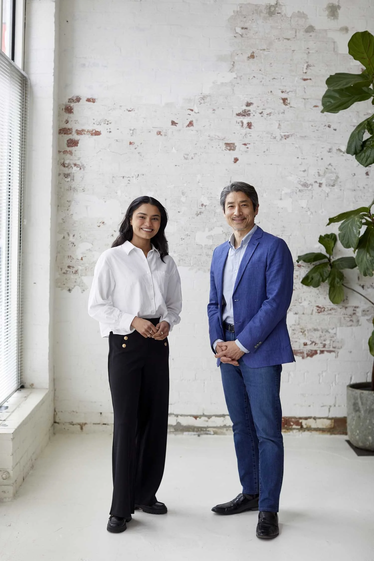 Two professionally dressed individuals, a young woman in a white shirt and black pants, and an older man in a blue blazer and jeans, standing side by side in a bright, minimalistic room with a white brick wall and large window to the left, smiling at