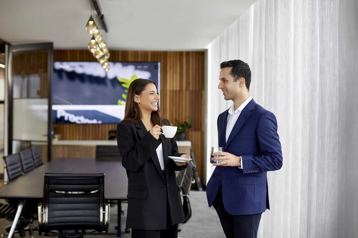 A woman and a man in business attire are talking and smiling in a modern office conference room. The woman is holding a cup and saucer, while the man holds a glass of water.