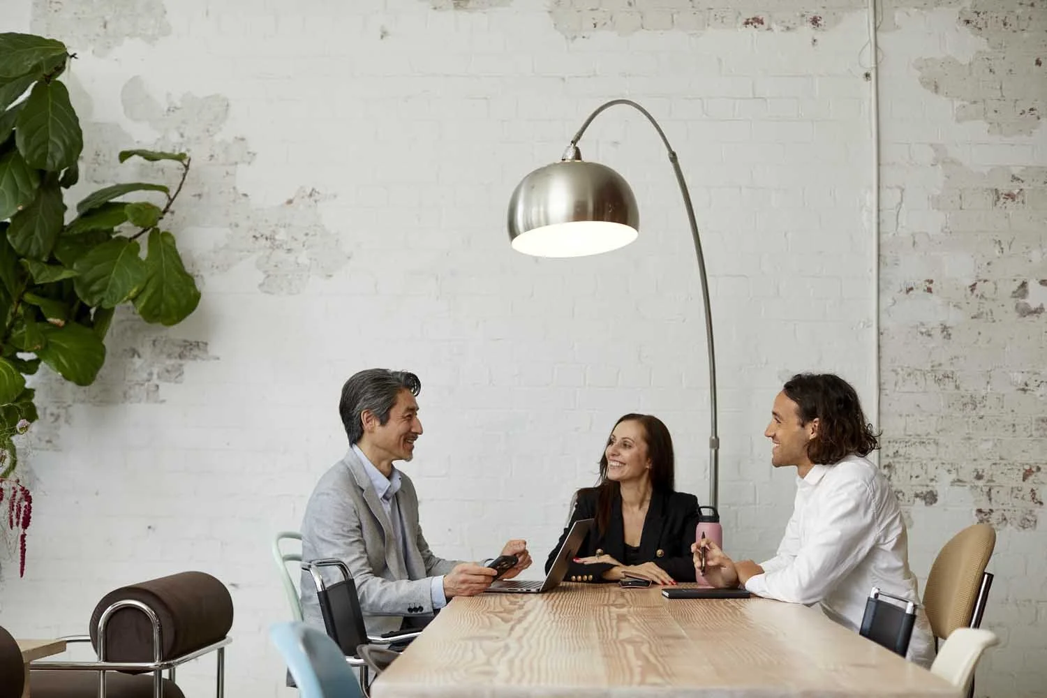 Three people having a meeting in a modern office space with a white brick wall, large green plant, and a tall, arched floor lamp.