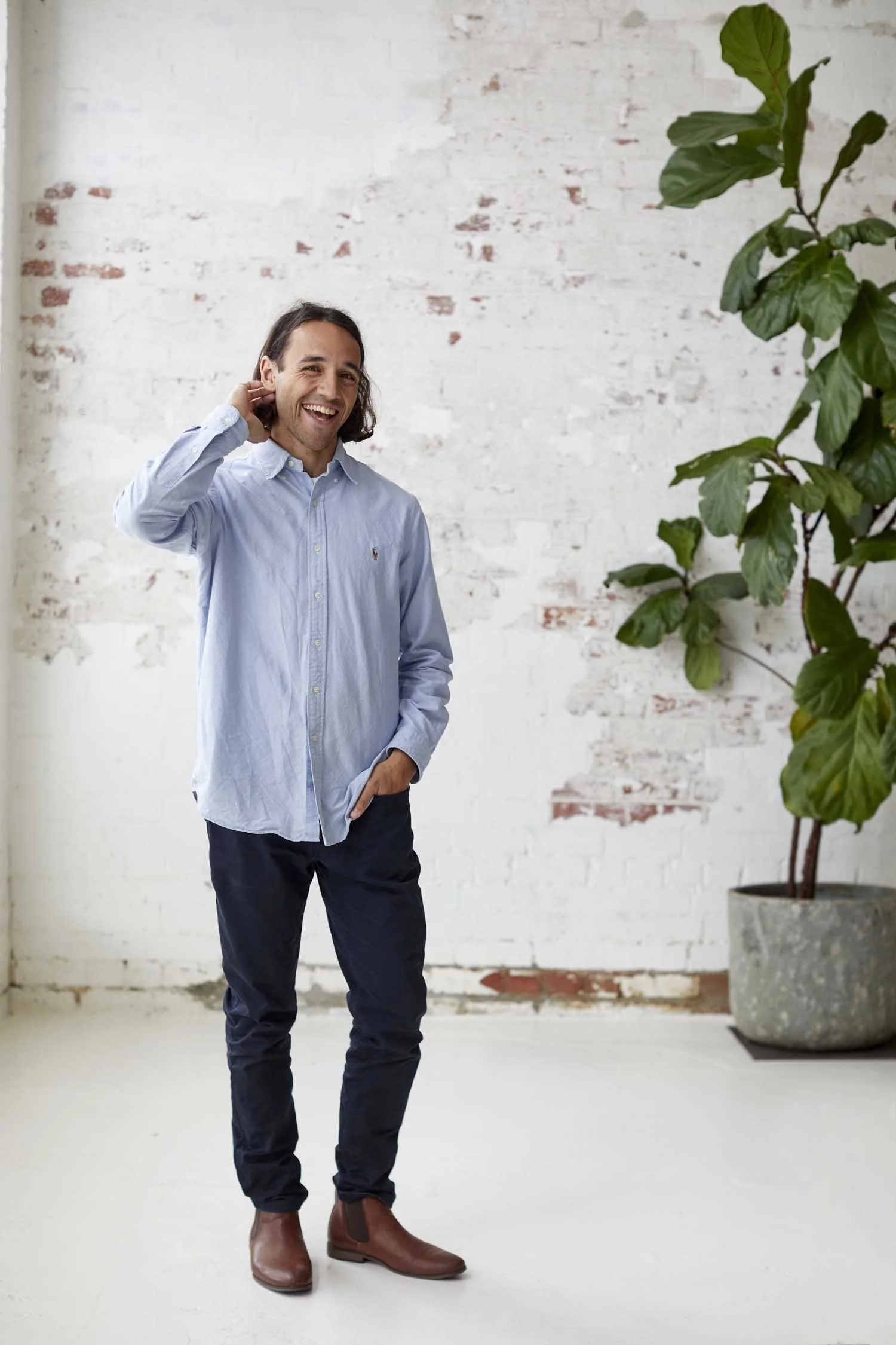 A man with long dark hair smiling and wearing a light blue button-up shirt, dark pants, and brown boots standing in front of a white brick wall with a large green potted plant on the right side.