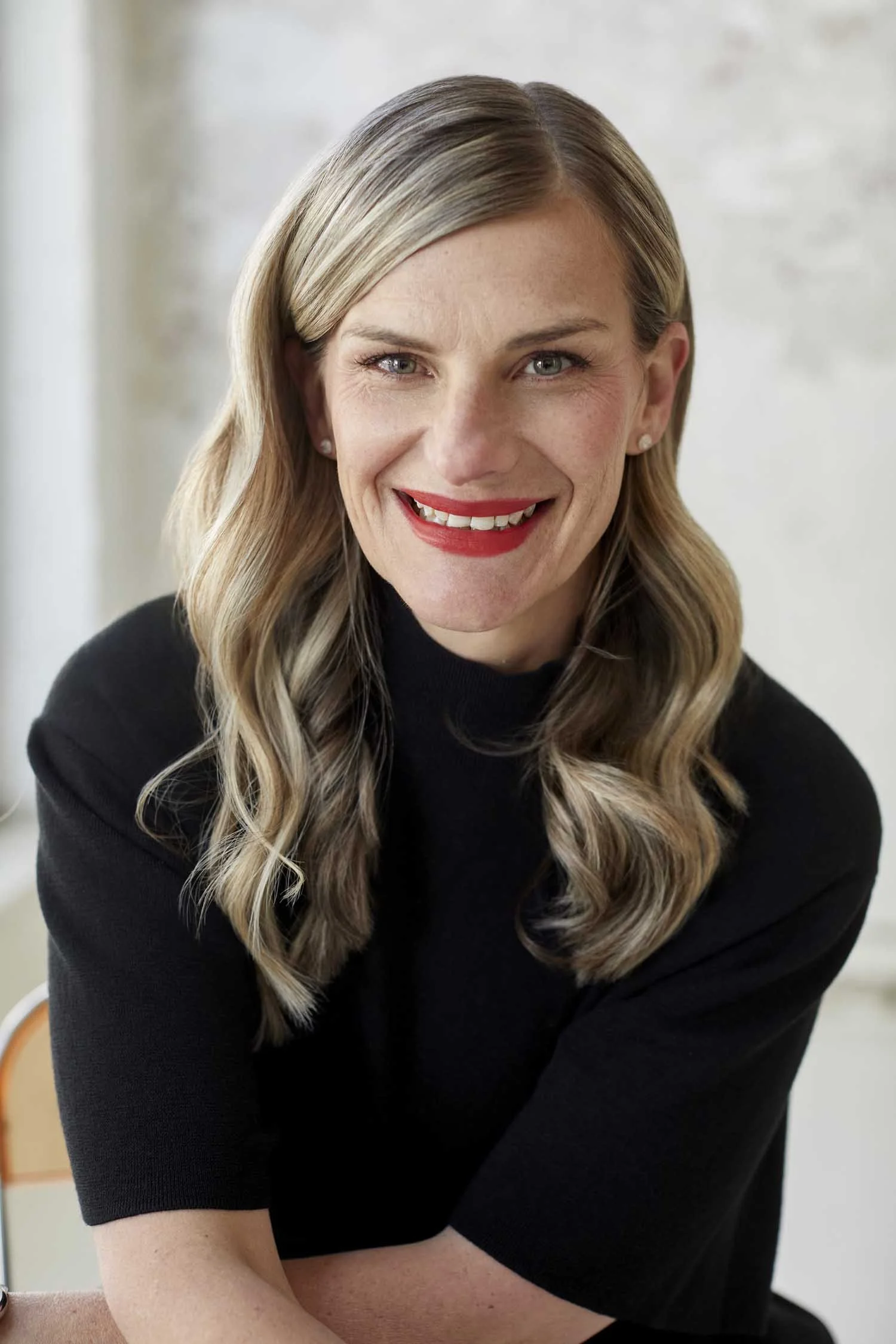 Close-up headshot of a woman with blonde hair, blue eyes, and red lipstick, wearing a black top, smiling, in an indoor setting.