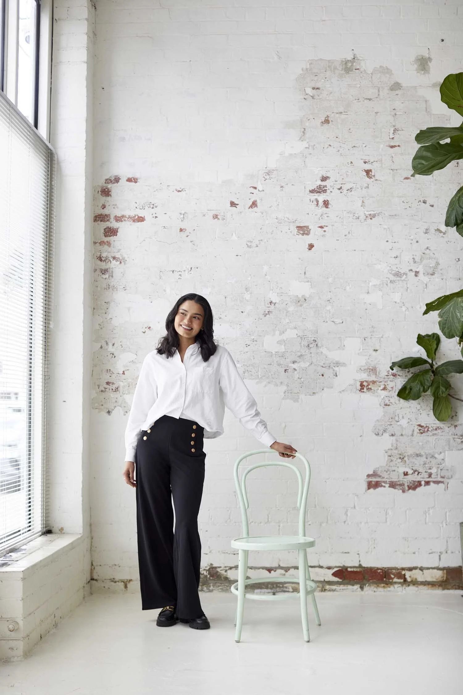 A woman with long dark hair standing in a bright room with a white brick wall. She is wearing a white blouse, black pants with gold buttons, and black shoes. She is smiling and touching a light green chair with her left hand. There is a large leafy plant on the right side of the image, and a window with blinds on the left.