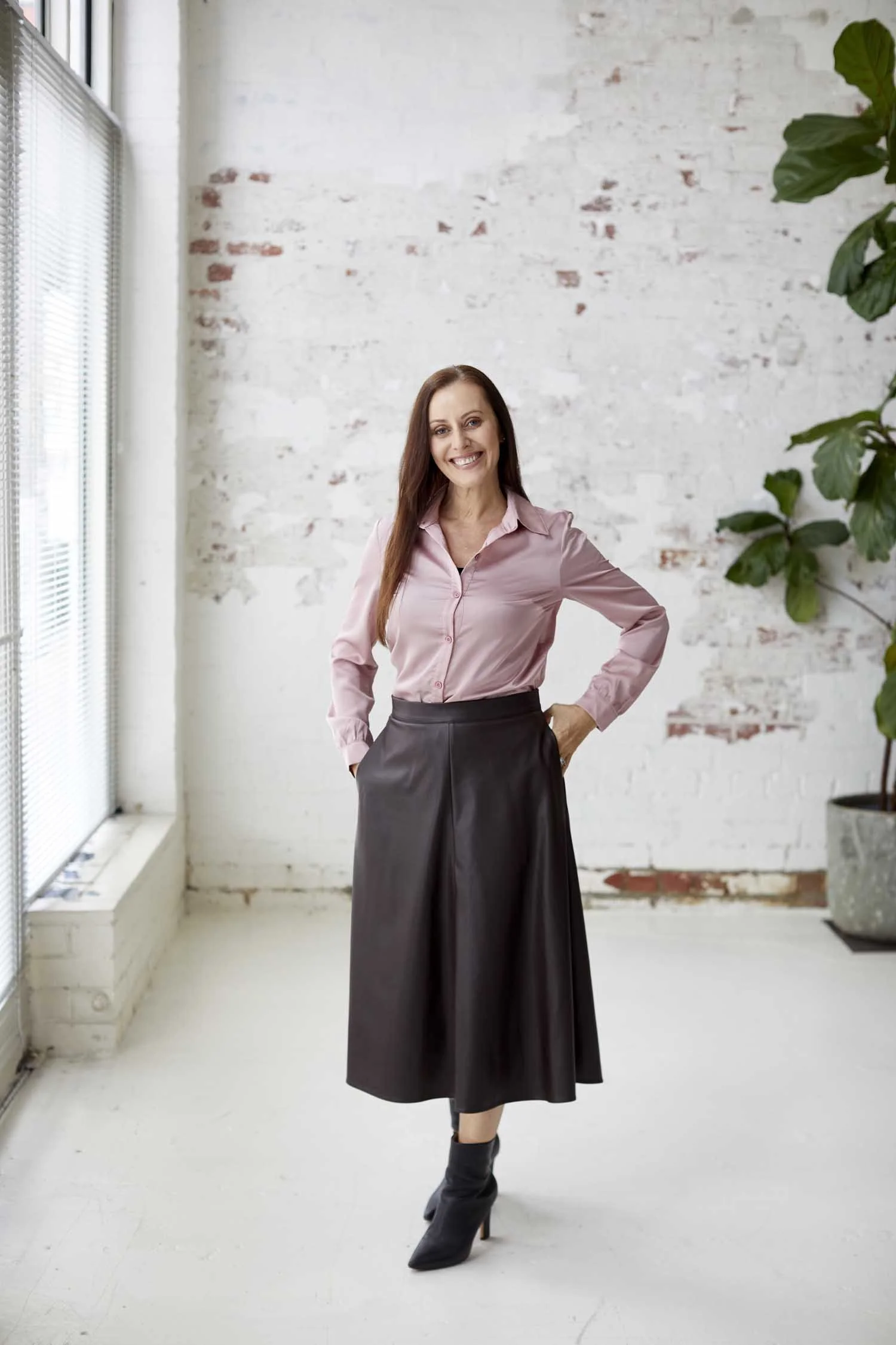 A woman with long hair and a smile standing in a bright, minimalist room with white brick walls, large windows with blinds, and a potted plant. She is wearing a pink button-up shirt and a black midi skirt, with one hand on her hip.