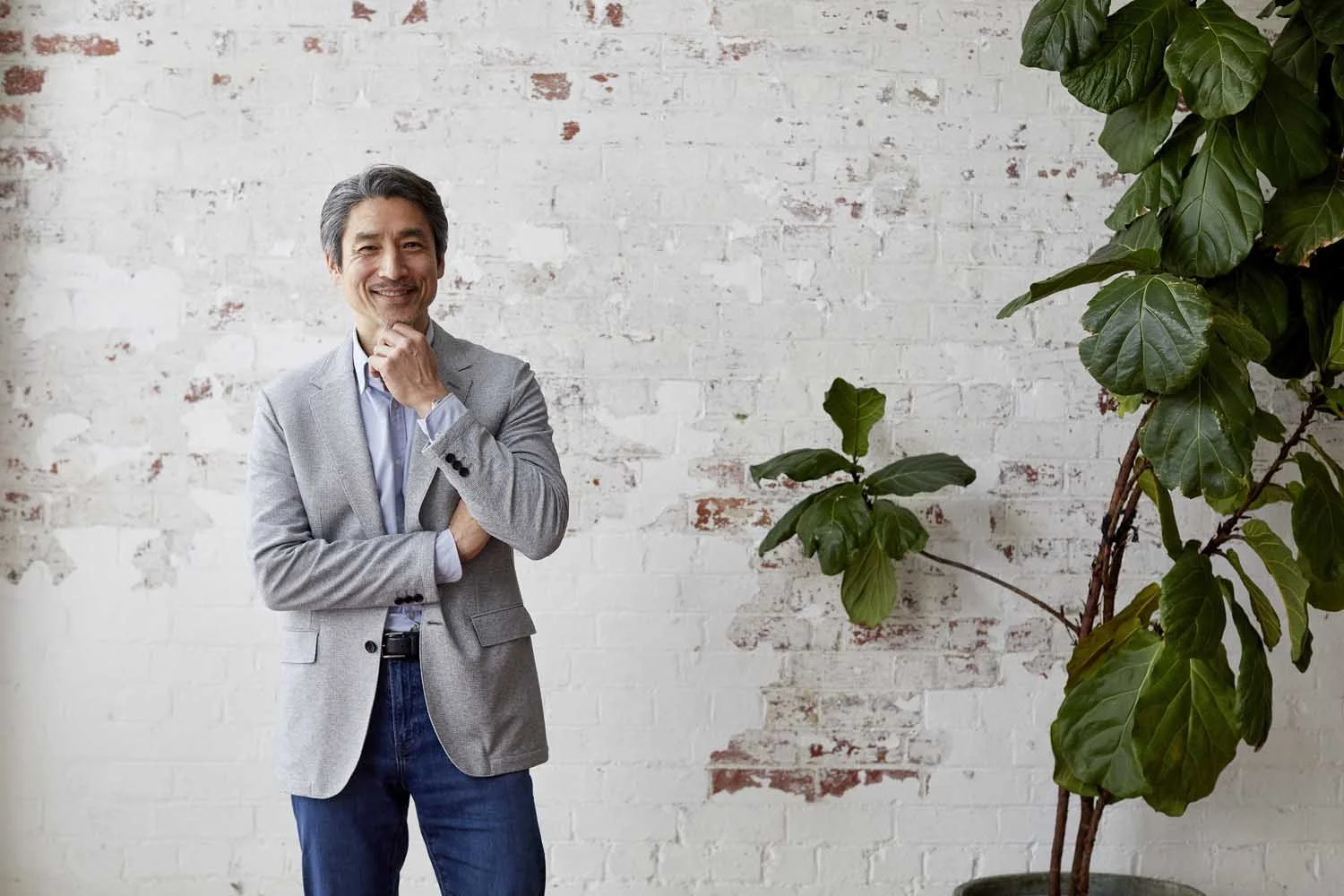 A middle-aged man in a gray blazer, light blue shirt, and jeans, smiling with his hand on his chin, standing near a large green houseplant against a white and brick wall.