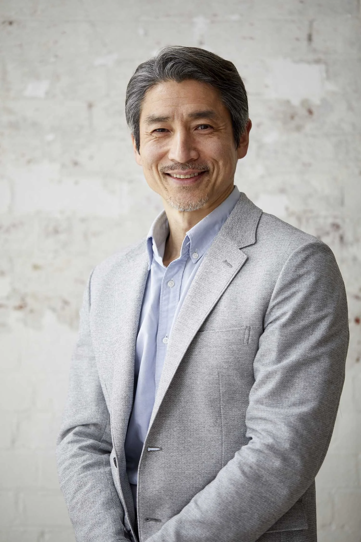 A middle-aged man in a light gray blazer and blue dress shirt smiling in front of a white brick wall.