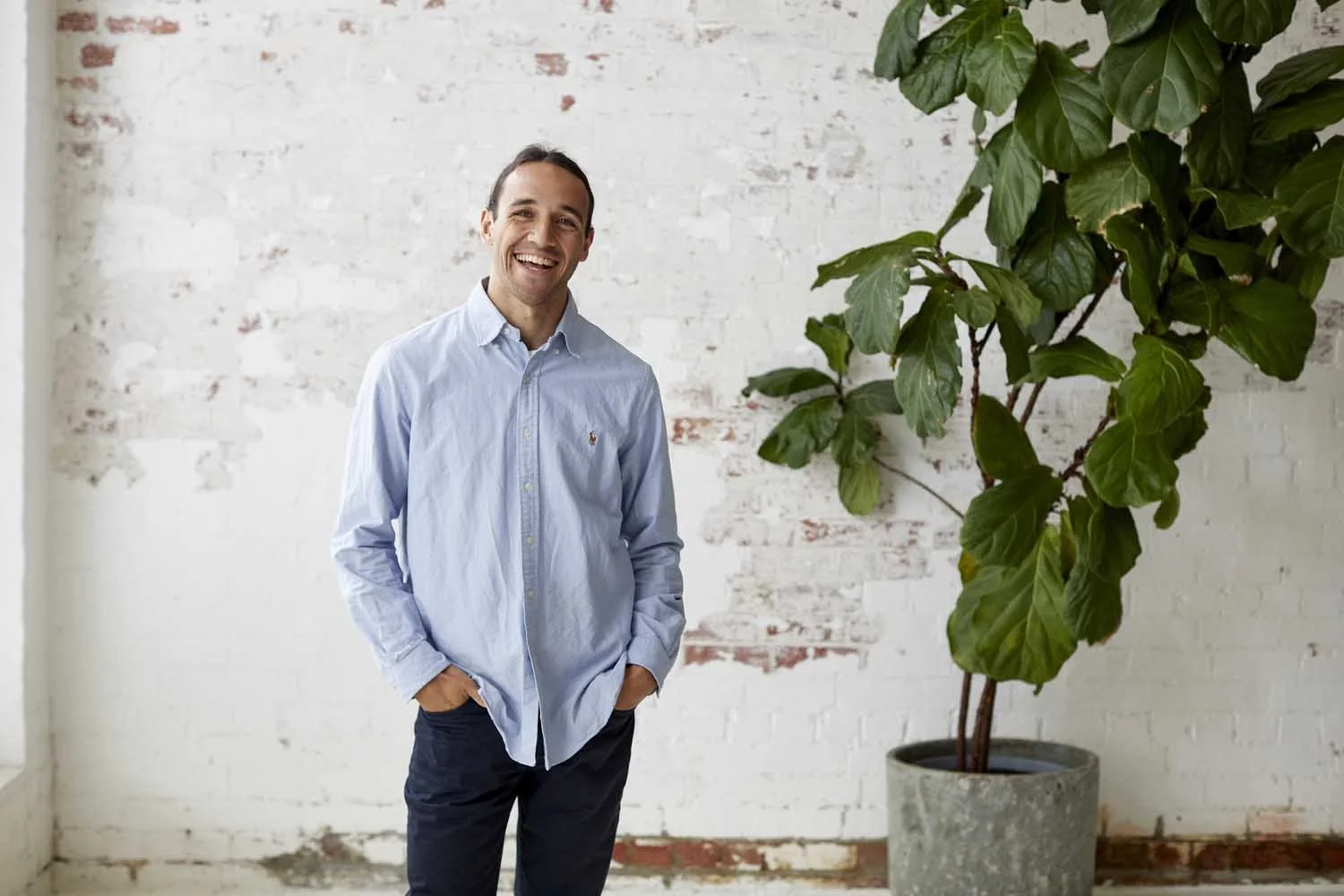 A man with dark hair in a light blue button-up shirt and dark pants smiling with his hands in his pockets, standing next to a large potted plant against a white brick wall.