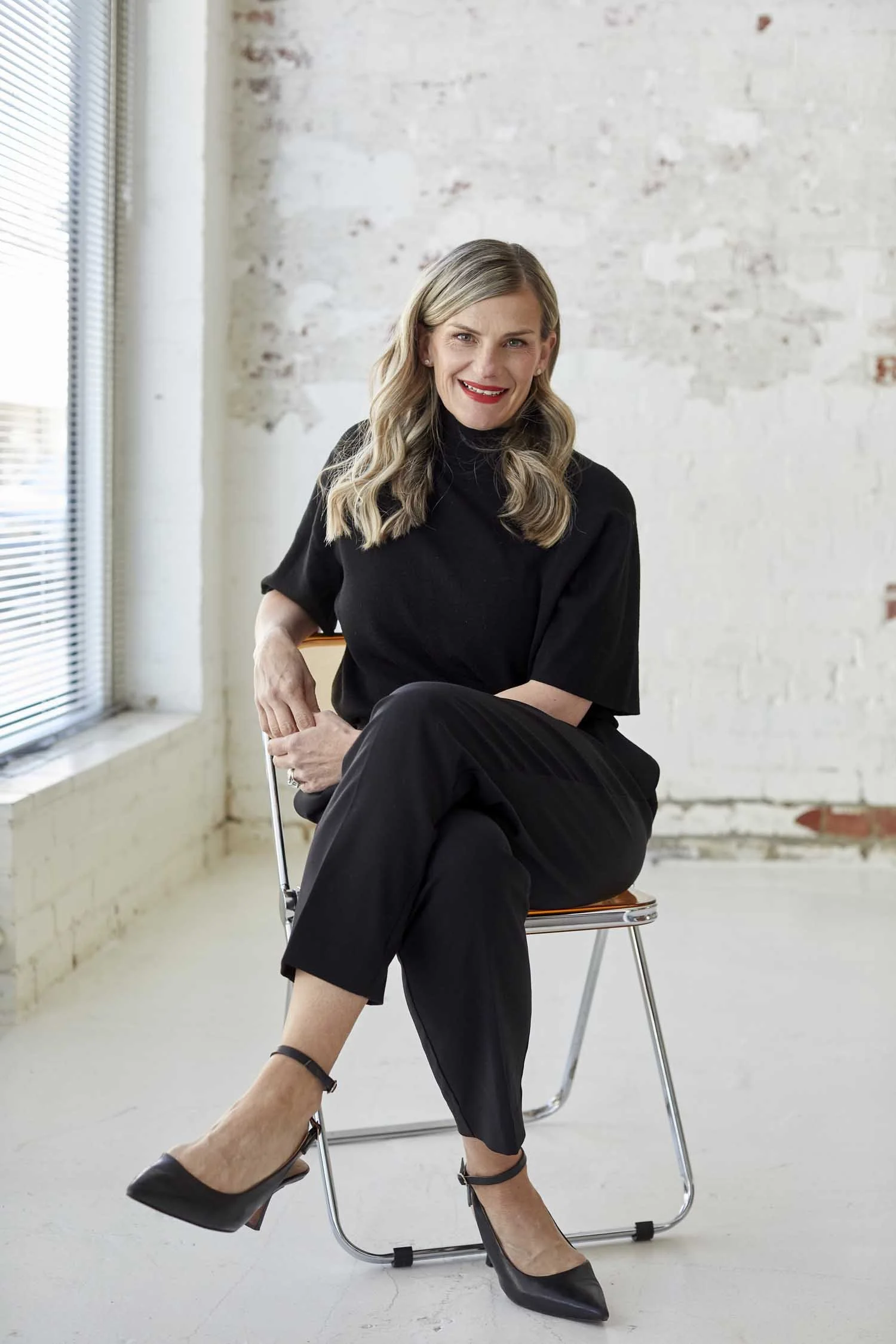 A woman with wavy blonde hair wearing a black top and black pants, sitting on a wooden chair in an industrial-style white brick room with large windows and blinds. She is smiling and looking at the camera, with one leg crossed over the other, and wea