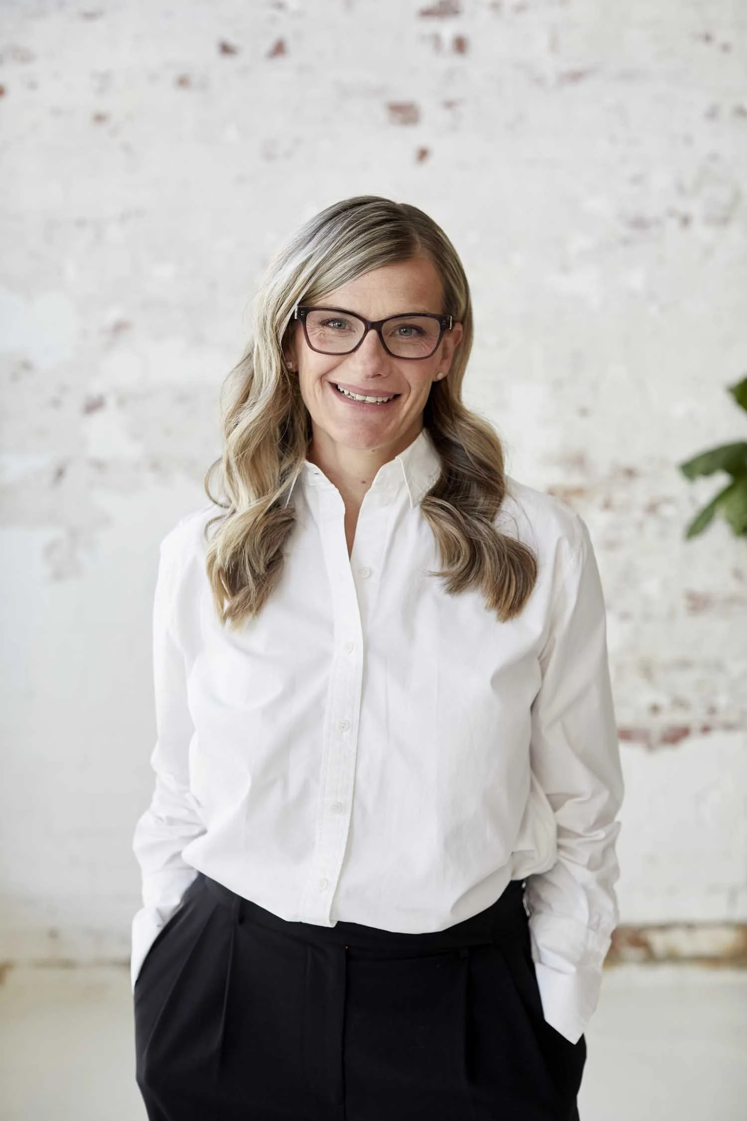 A woman with wavy blond hair, wearing glasses, a white blouse, and black pants, standing in front of a white brick wall with some greenery visible on the right side, smiling at the camera.