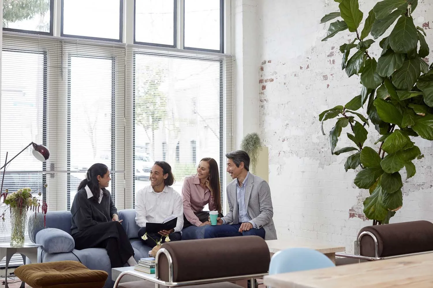 Four people sitting on a blue couch in a modern office space, engaged in conversation and smiling, with large windows and indoor plants around them.