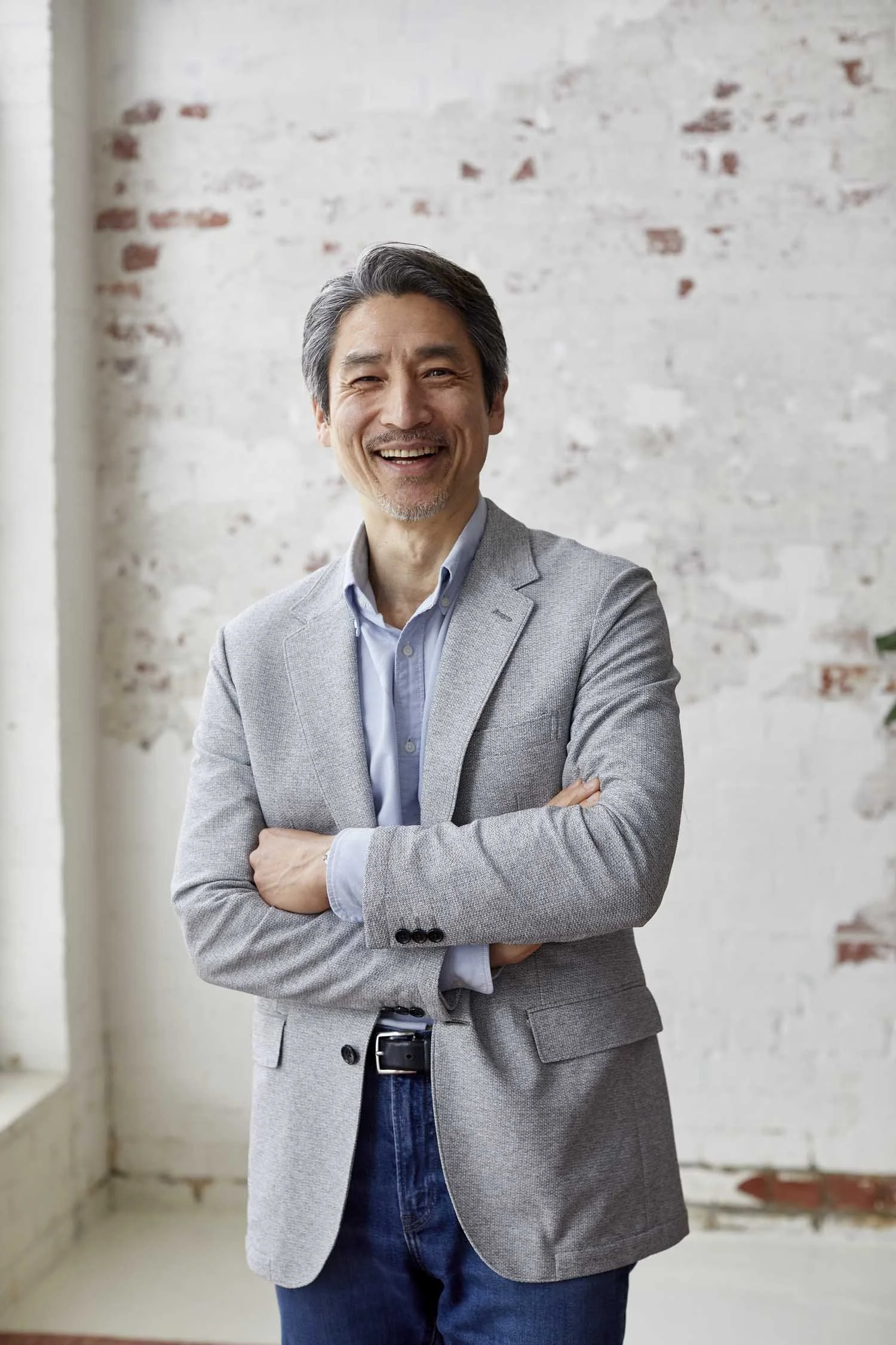 A smiling man with gray hair and a beard, wearing a light gray blazer, blue shirt, and jeans, standing with arms crossed in front of a white brick wall.
