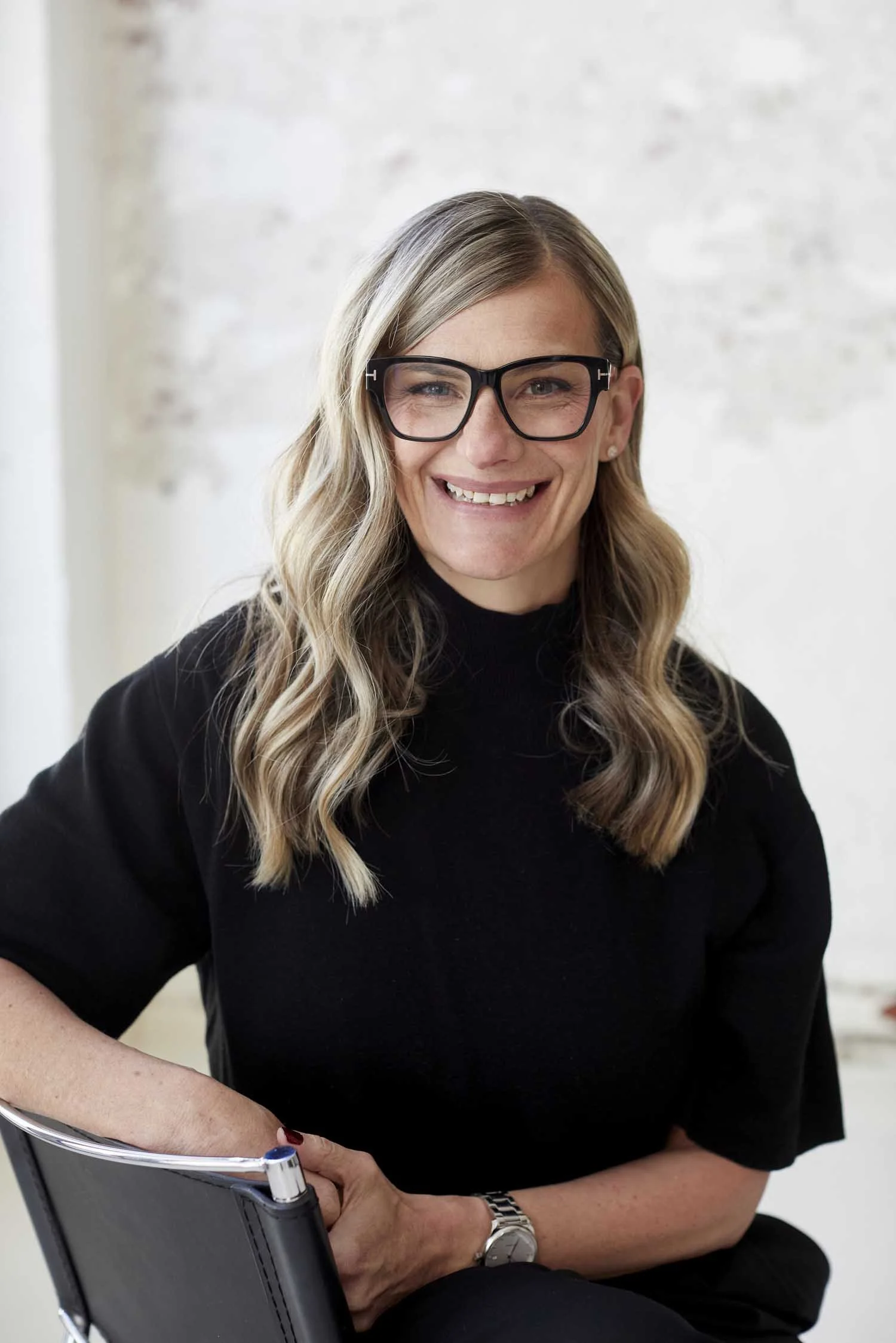 A woman with long blonde hair, glasses, and a black top sitting in a chair, smiling, with a white wall background.