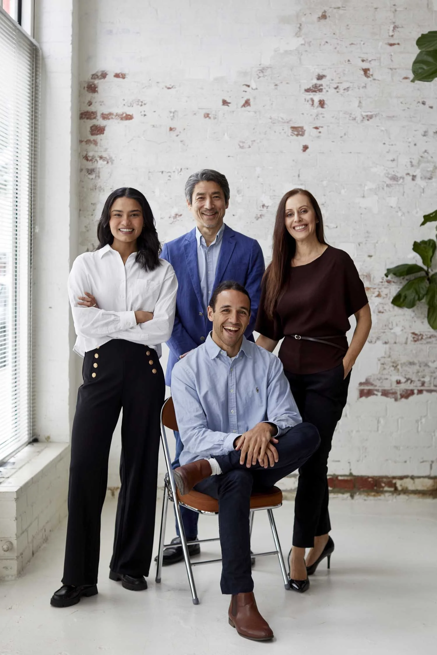 Group of five diverse professionals posing together in a bright modern office with white brick wall, large window with blinds, and some green plants. They are smiling and dressed in business casual attire.