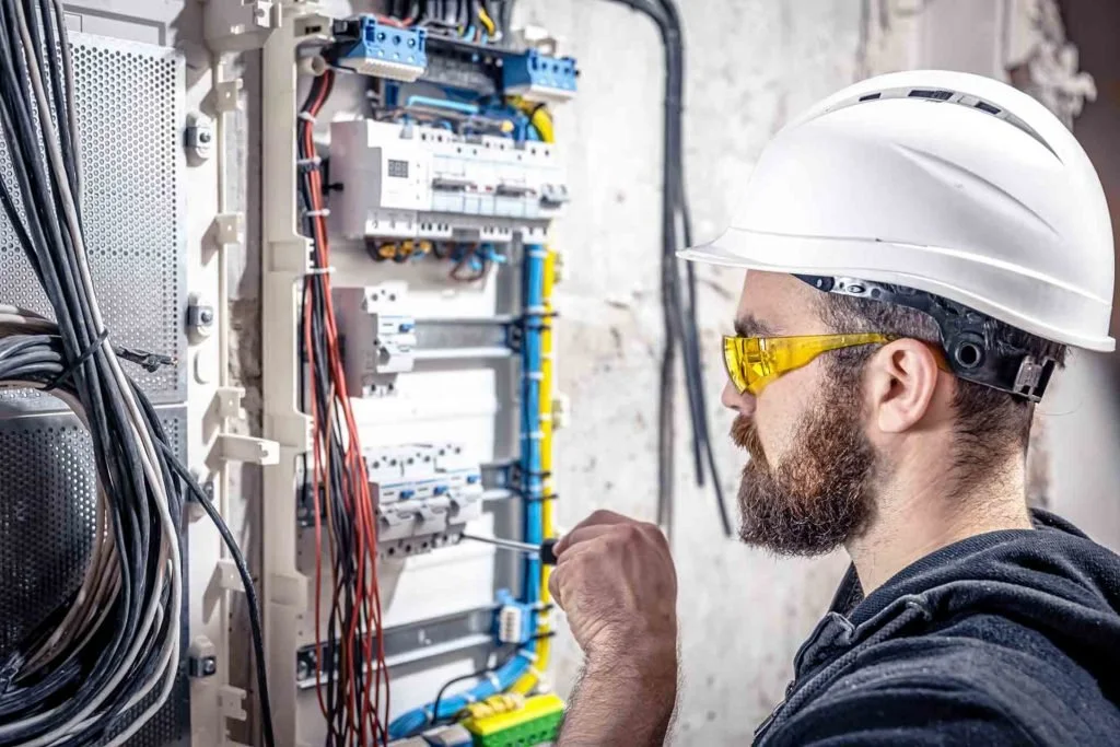 A male electrician wearing a white safety helmet and yellow safety glasses working on an electrical panel with various wires and components.