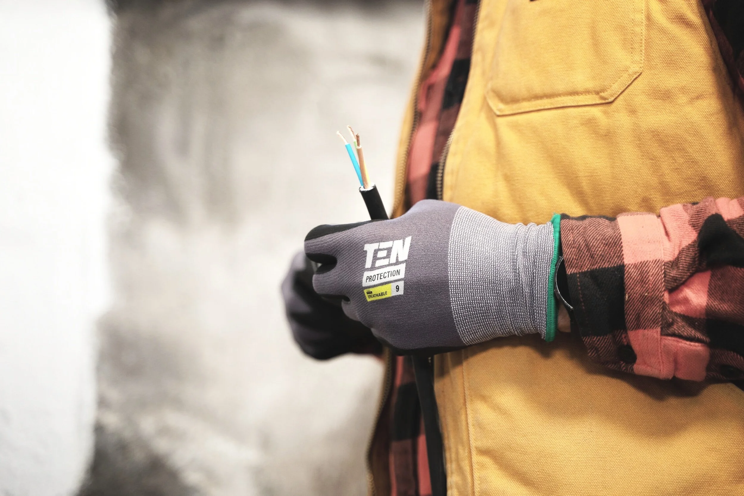 A person wearing a gray protective glove holding a black cable with multiple colored wires exposed, standing beside a yellow jacket and a pink and black plaid shirt.