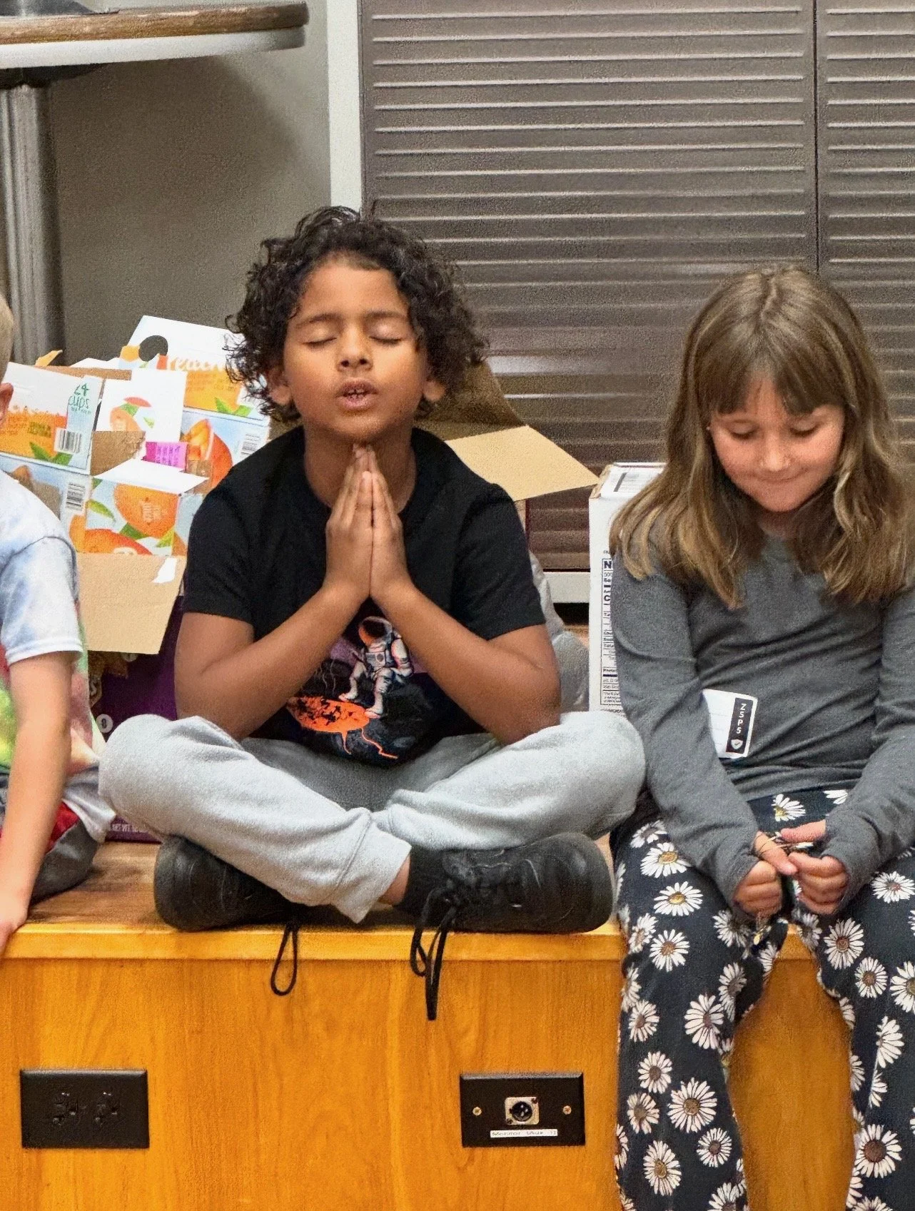 Children sitting cross-legged on a wooden stage, with their hands in prayer position, eyes closed. Behind them are cardboard boxes and a wooden backdrop.