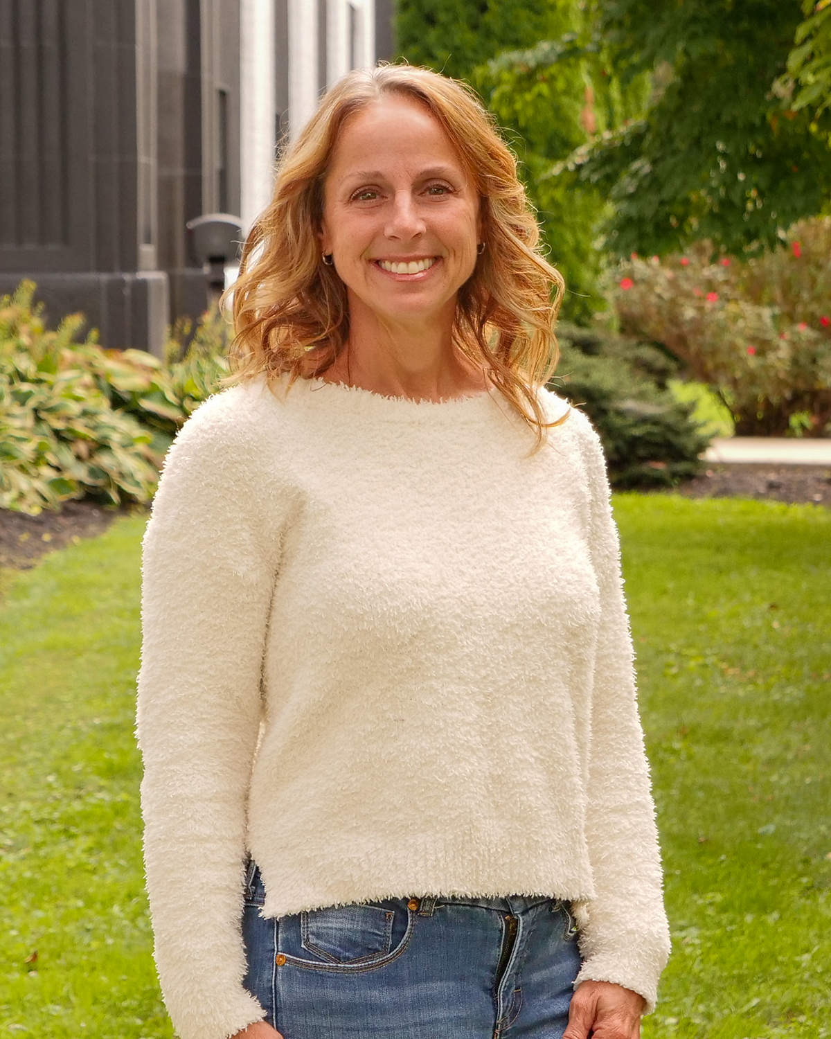 A woman with curly blonde hair smiling outdoors, wearing a sleeveless white dress with beige accents, standing on a grassy lawn with trees and a building in the background.