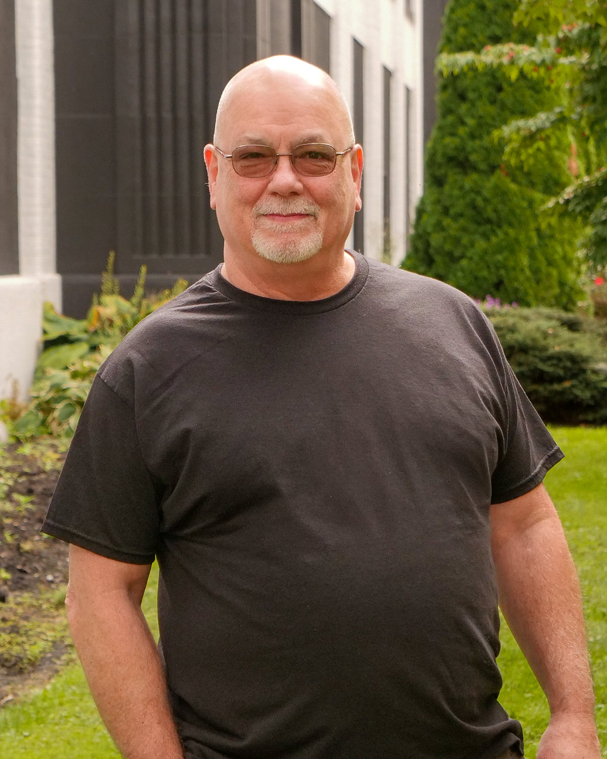 A smiling man with glasses and a beard standing outdoors in a yard with green grass and trees, next to a building with large windows.