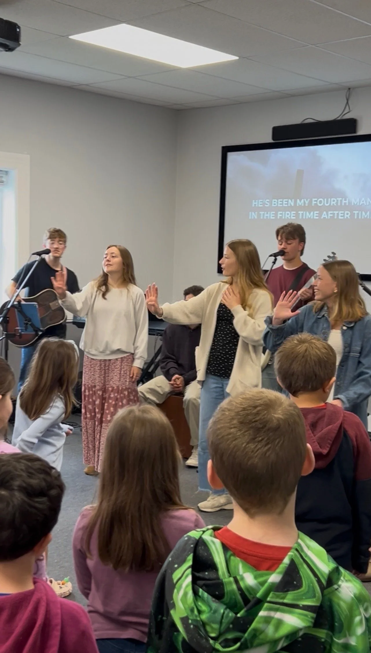 A group of young people and children inside a classroom or worship space, participating in a musical or religious activity. Some are singing or gesturing, with one person playing guitar. The background features a large screen displaying song lyrics.