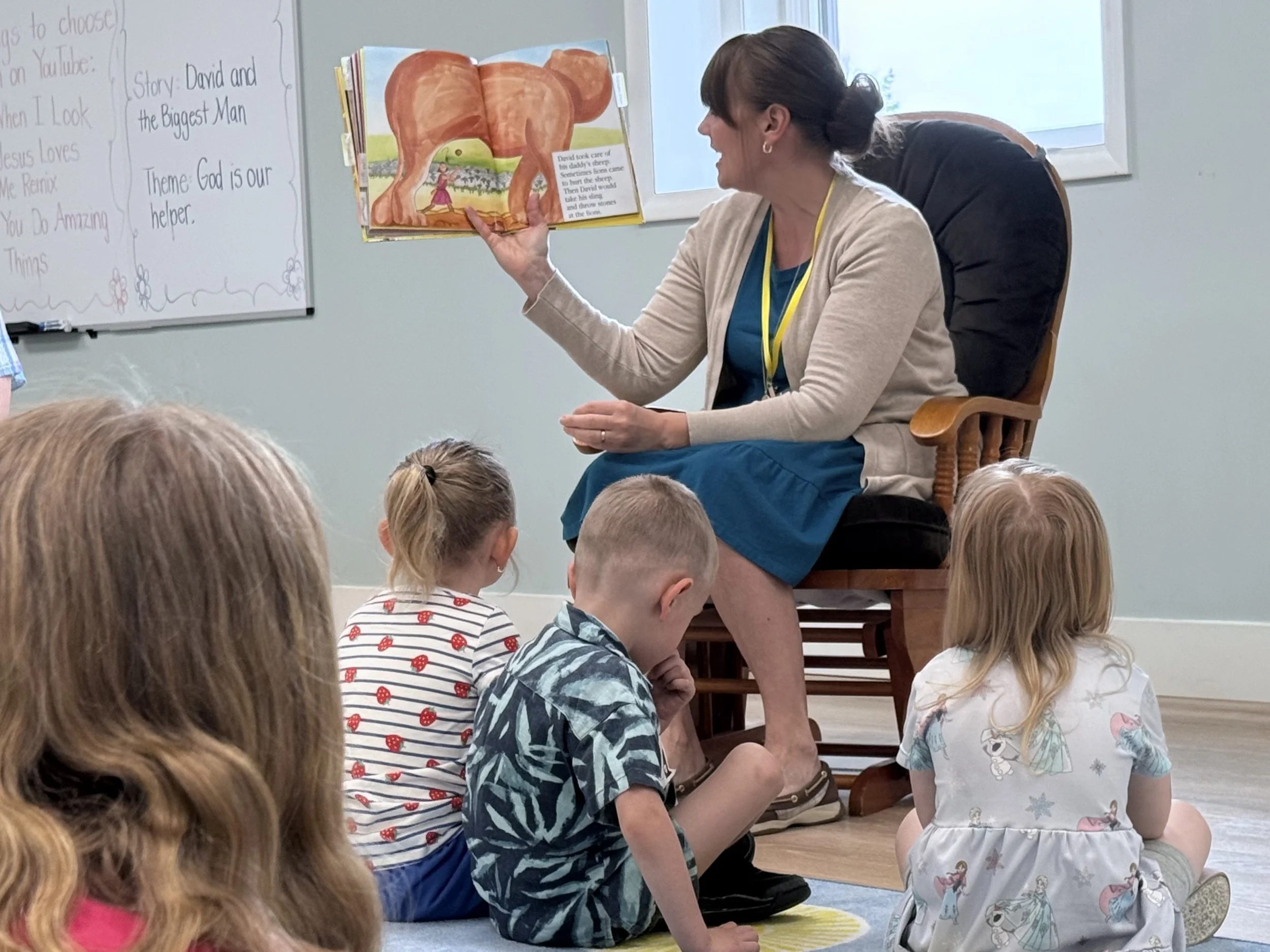 A woman sitting on a wooden chair in front of a group of children, reading a colorful picture book with an illustration of a large dog and a girl. The children are sitting on the floor, listening attentively. There is a whiteboard with writing behind her.