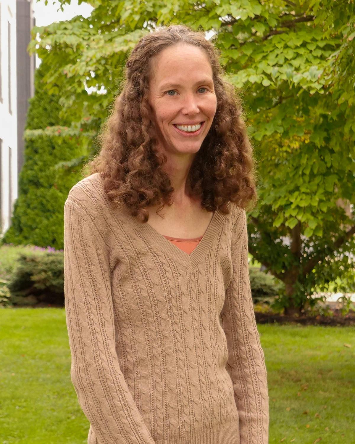 A woman with long curly hair, wearing a black shirt and checkered pants, standing outdoors in front of a large green bush, smiling at the camera.