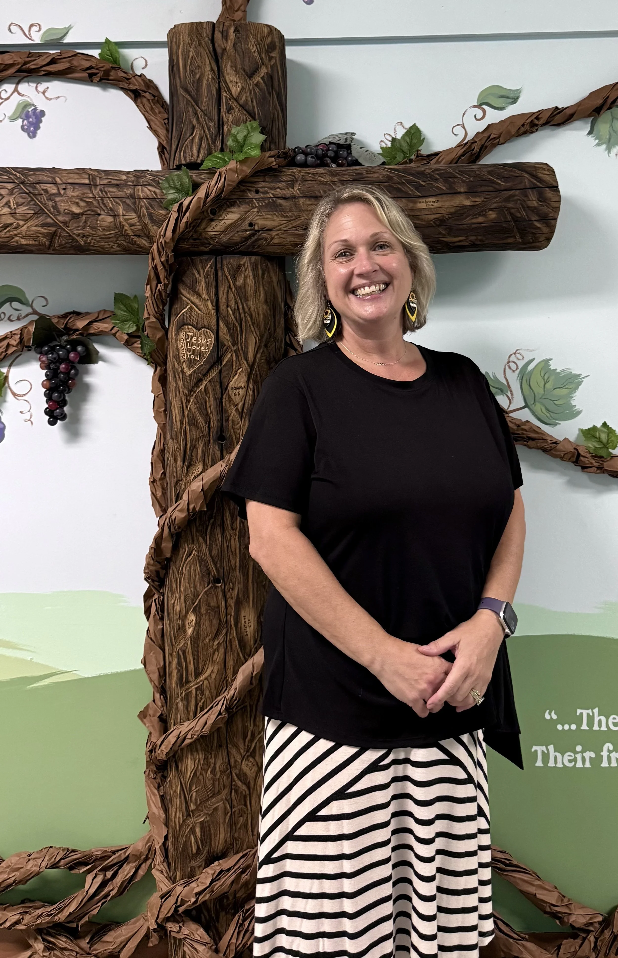 A smiling woman with blonde hair, wearing black and white striped pants, a black top, earrings, and a smartwatch, standing in front of a wooden cross decorated with vines, leaves, and grapes, against a painted background.