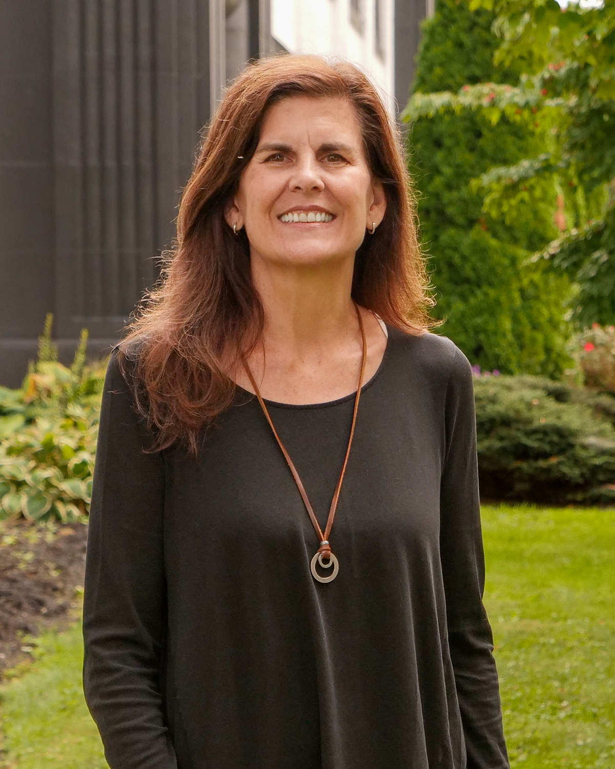 A middle-aged woman with long brown hair standing outdoors in front of a modern building, smiling, wearing a white sleeveless dress with black line patterns, with green foliage and shrubs around her.
