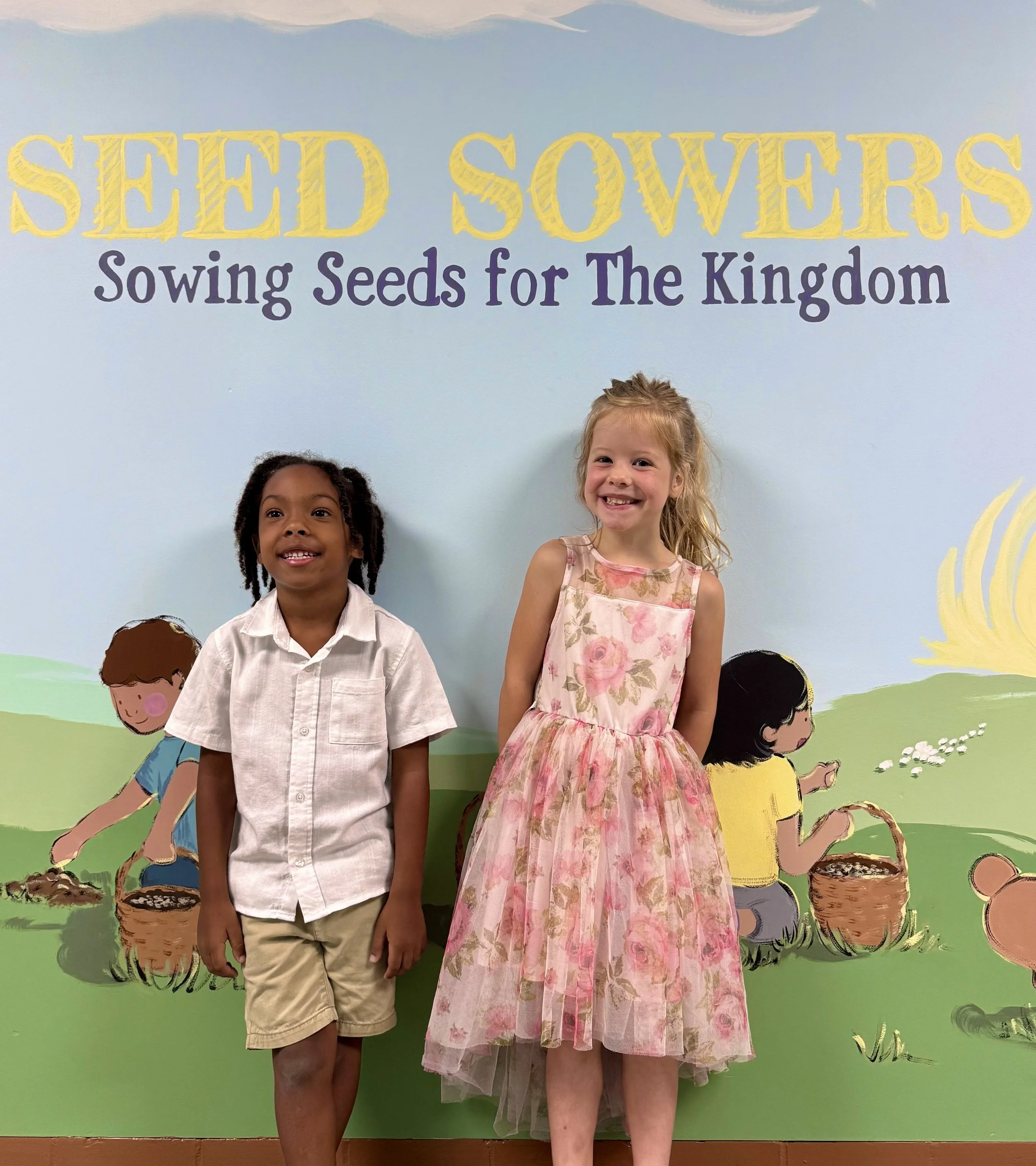 Two young girls standing in front of a wall with colorful cartoon illustrations of children planting seeds, with the phrase 'Seed Sowears: Sowing Seeds for The Kingdom' written above in decorative yellow and purple letters.