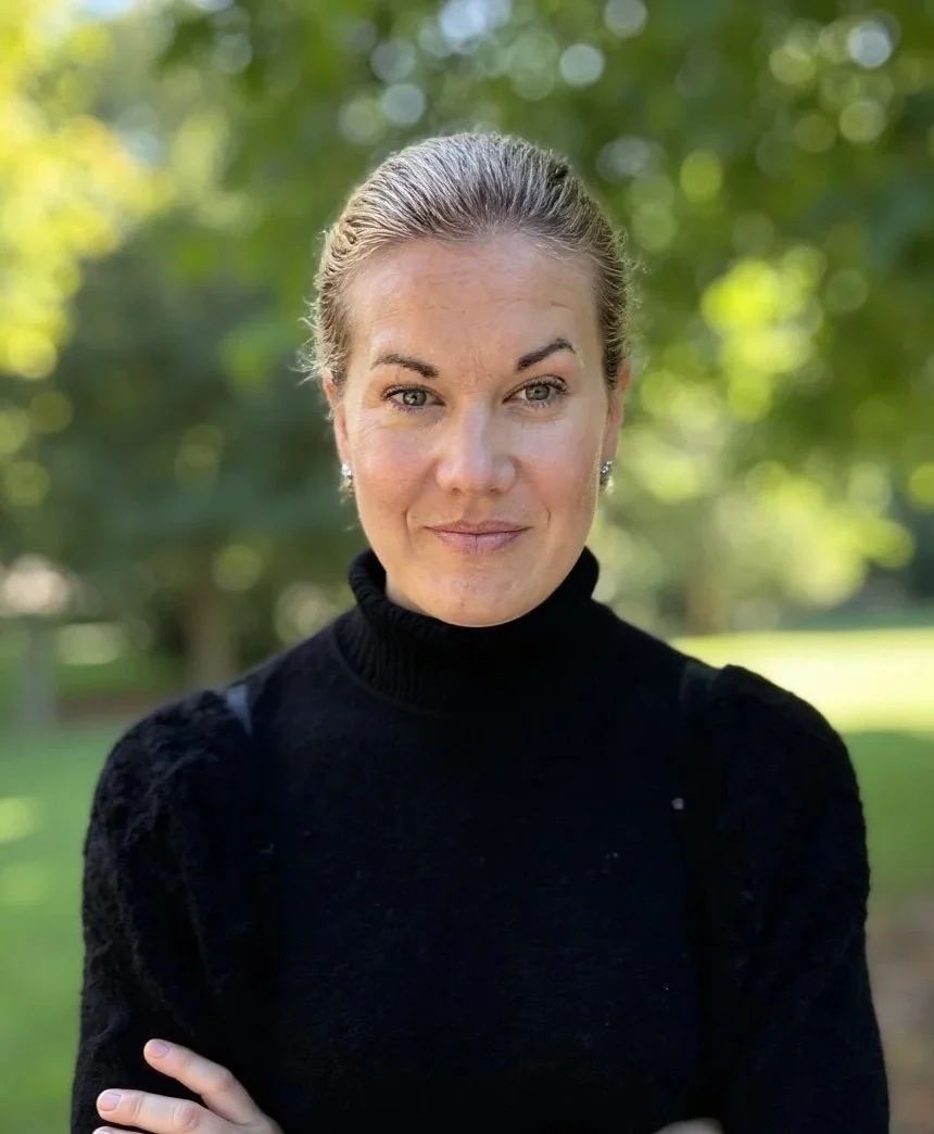 Porträtt av en kvinna med ljus hud, ljusbrunt hår i uppsättning, som bär svart turtleneck-tröja, utomhus med gröna träd i bakgrunden, leende.