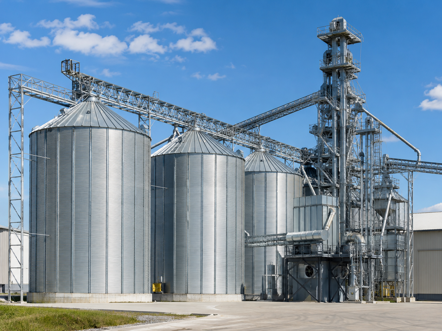 Four large metallic grain silos with a red conveyor system above them, set against a partly cloudy sky.